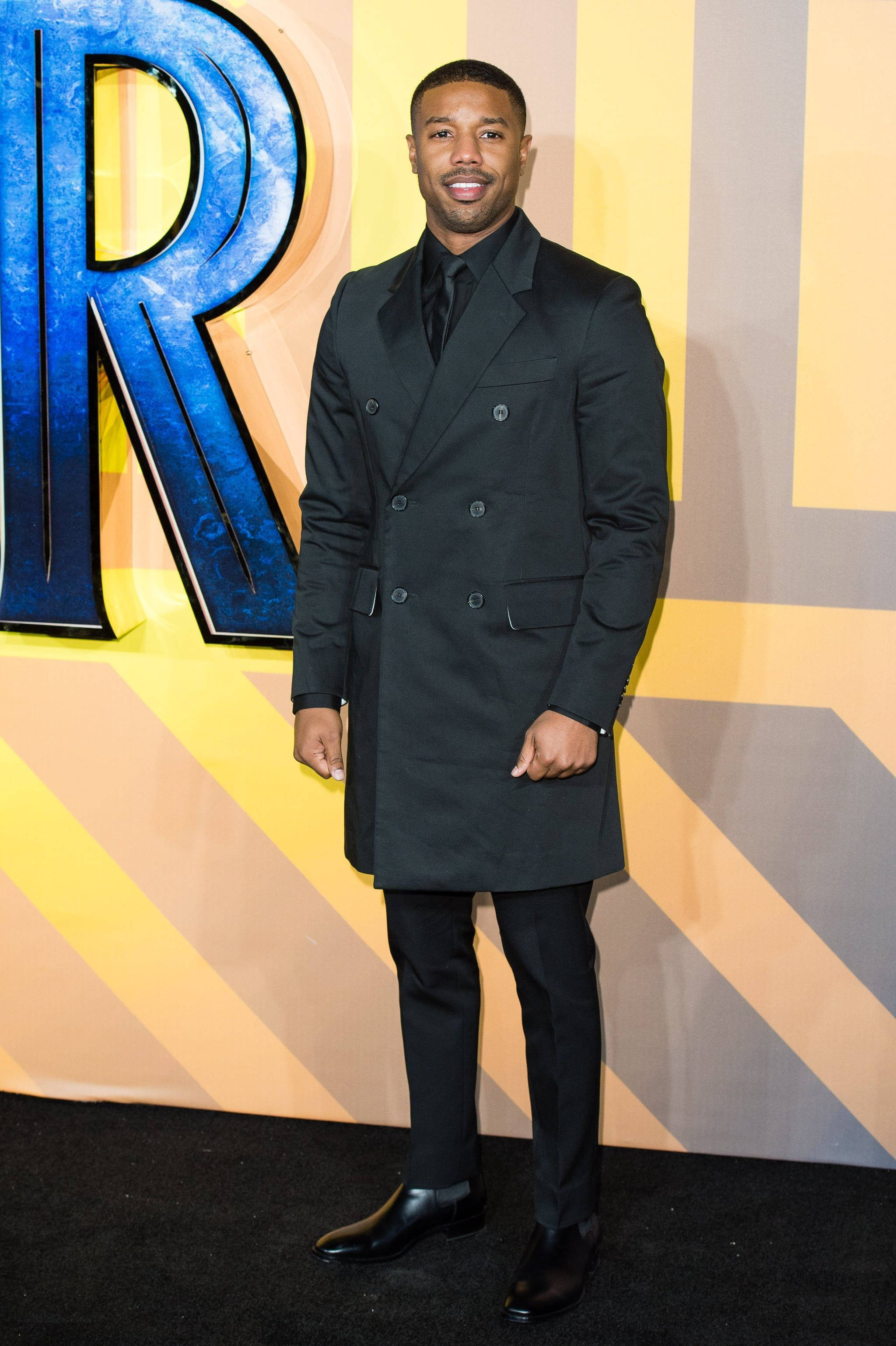 michael 桌布 壁紙 — 演員 Michael B Jordan 在充滿活力的活動背景下，穿著俐落的黑色雙排扣大衣自信地微笑。