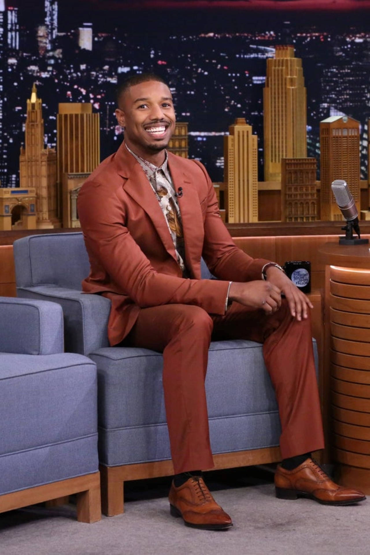 michael 桌布 壁紙 — 演員 Michael B Jordan 穿著鏽色西裝，在脫口秀佈景中溫暖微笑，背景是閃爍的城市天際線。