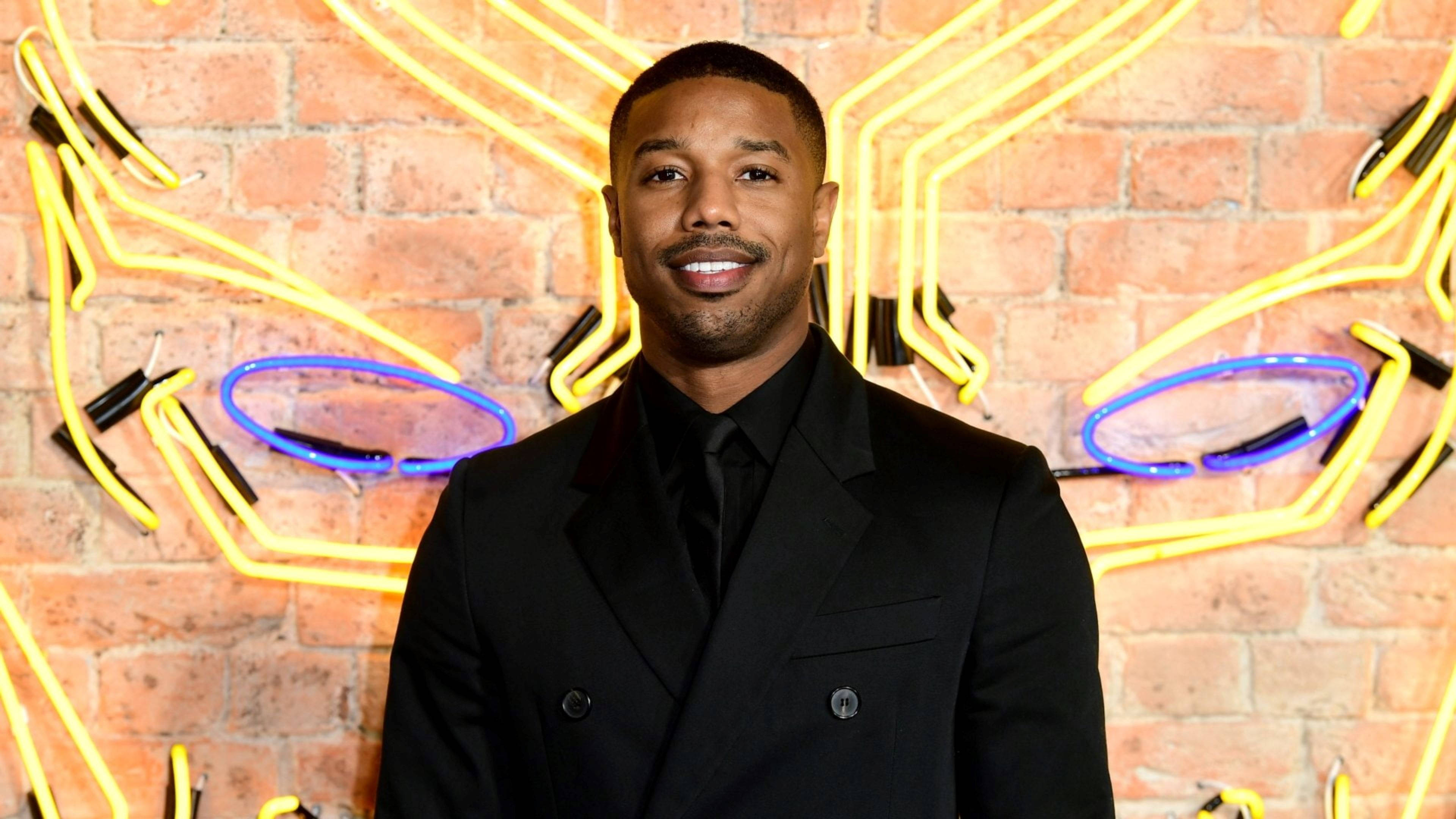 michael 桌布 壁紙 — 演員 Michael B Jordan 自信地微笑，身穿黑色西裝，背景是裝飾著黃色和藍色霓虹燈的磚牆。