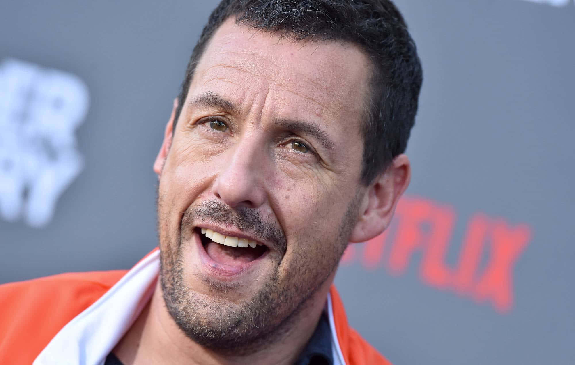 Adam Sandler 桌布 壁紙 — 演員亞當·山德勒在Netflix活動中穿著橙色外套，露出燦爛的笑容。