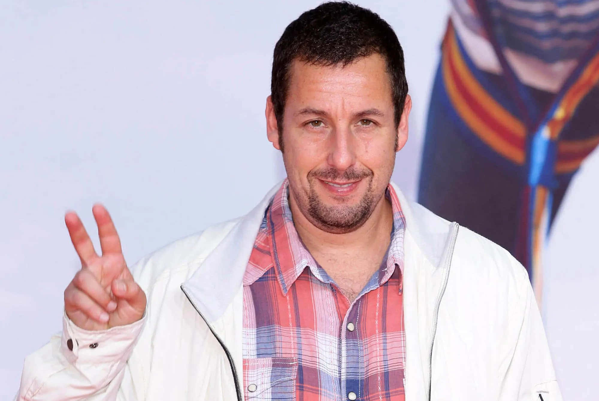 Adam Sandler 桌布 壁紙 — 演員亞當·山德勒穿著白色外套和格紋襯衫，對著鏡頭溫暖微笑，同時做出和平手勢。