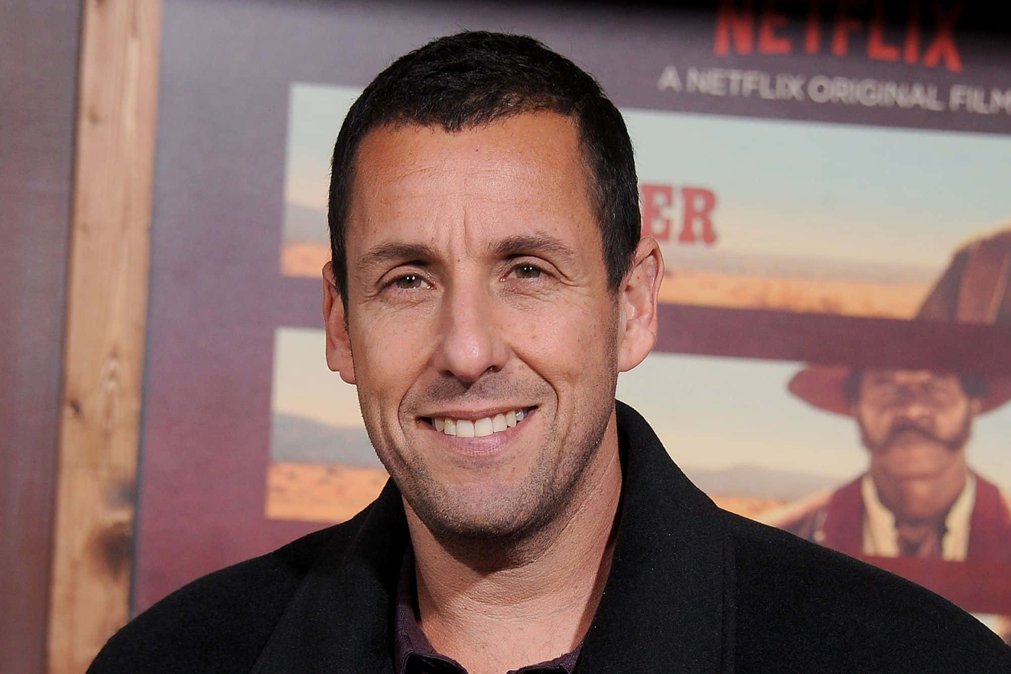 Adam Sandler 桌布 壁紙 — 演員亞當·山德勒穿著黑色外套，在Netflix電影海報背景下溫暖地對著鏡頭微笑。
