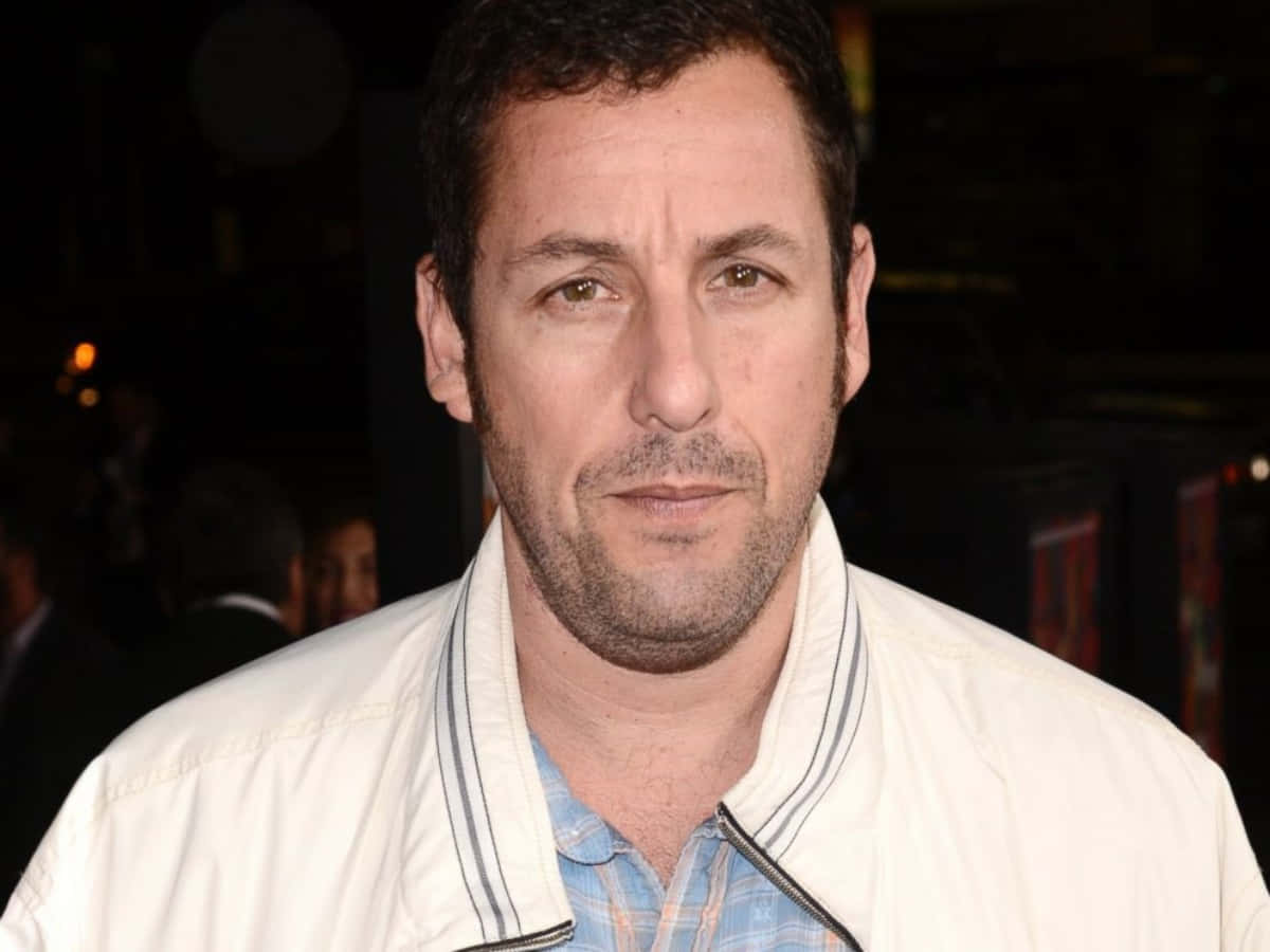 Adam Sandler 桌布 壁紙 — 演員亞當·山德勒身穿白色外套和藍色格紋襯衫，在夜間活動中，背景燈光模糊。