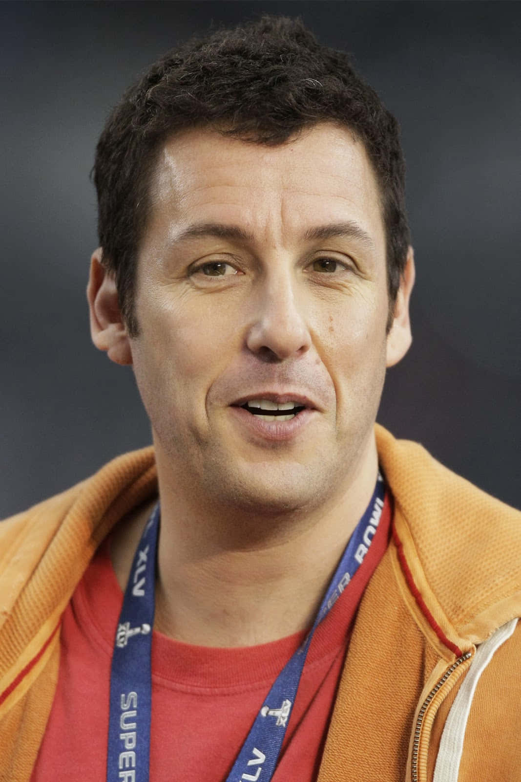 Adam Sandler 桌布 壁紙 — 演員亞當·桑德勒的特寫，穿著橙色連帽衫和超級碗掛繩，背景為深色。
