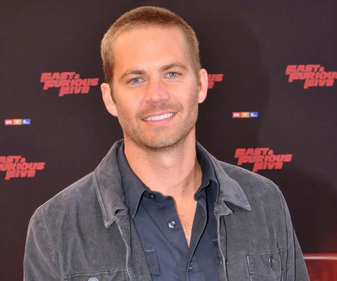 Paul Walker 桌布 壁紙 — 演員保羅·沃克穿著灰色燈芯絨外套內搭深色襯衫，在《玩命關頭5》宣傳背景前對著鏡頭溫暖微笑。