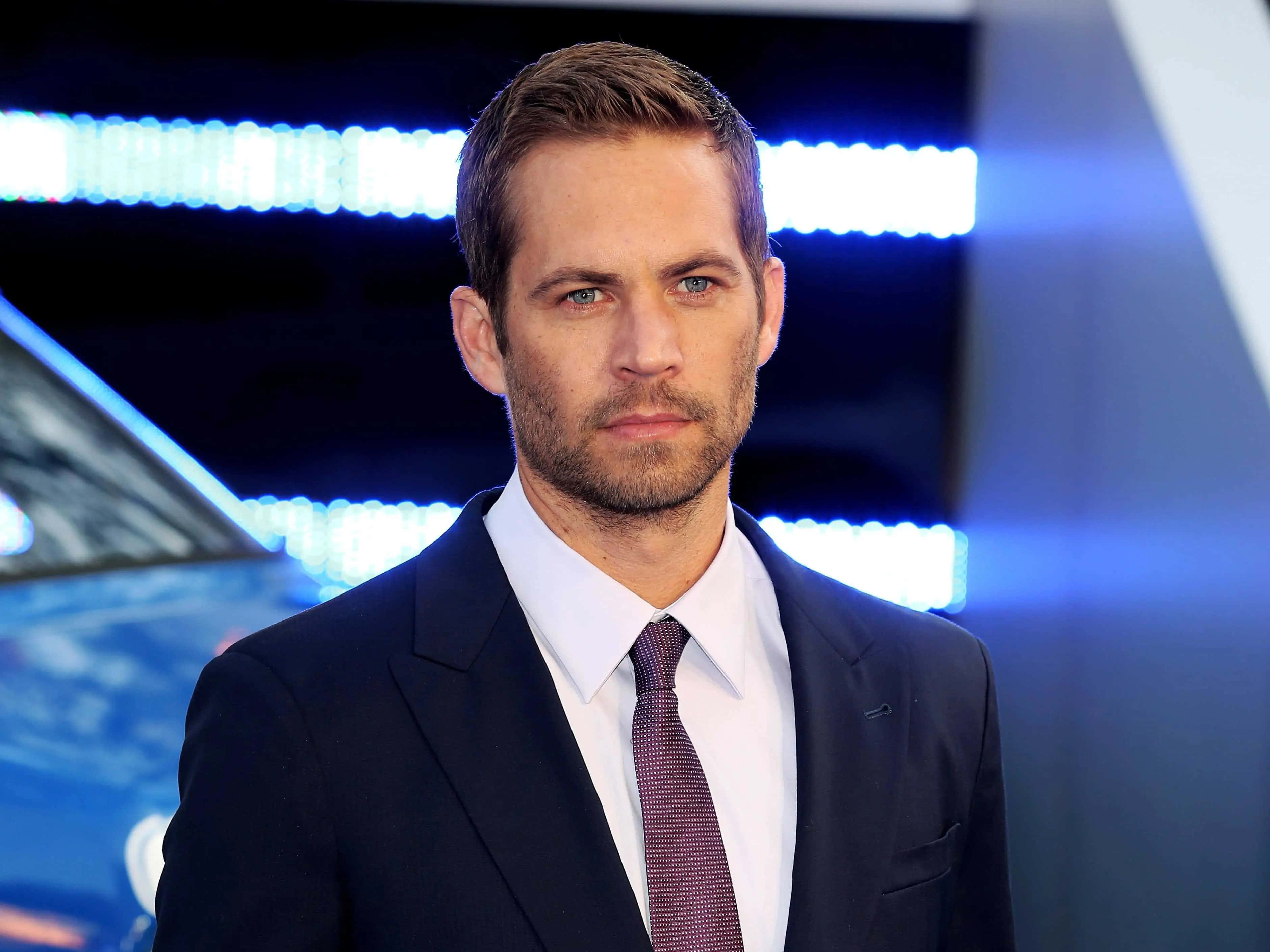 Paul Walker 桌布 壁紙 — 演員保羅·沃克身穿深色西裝和領帶，自信地站在藍色燈光背景前，身後可見一輛汽車。