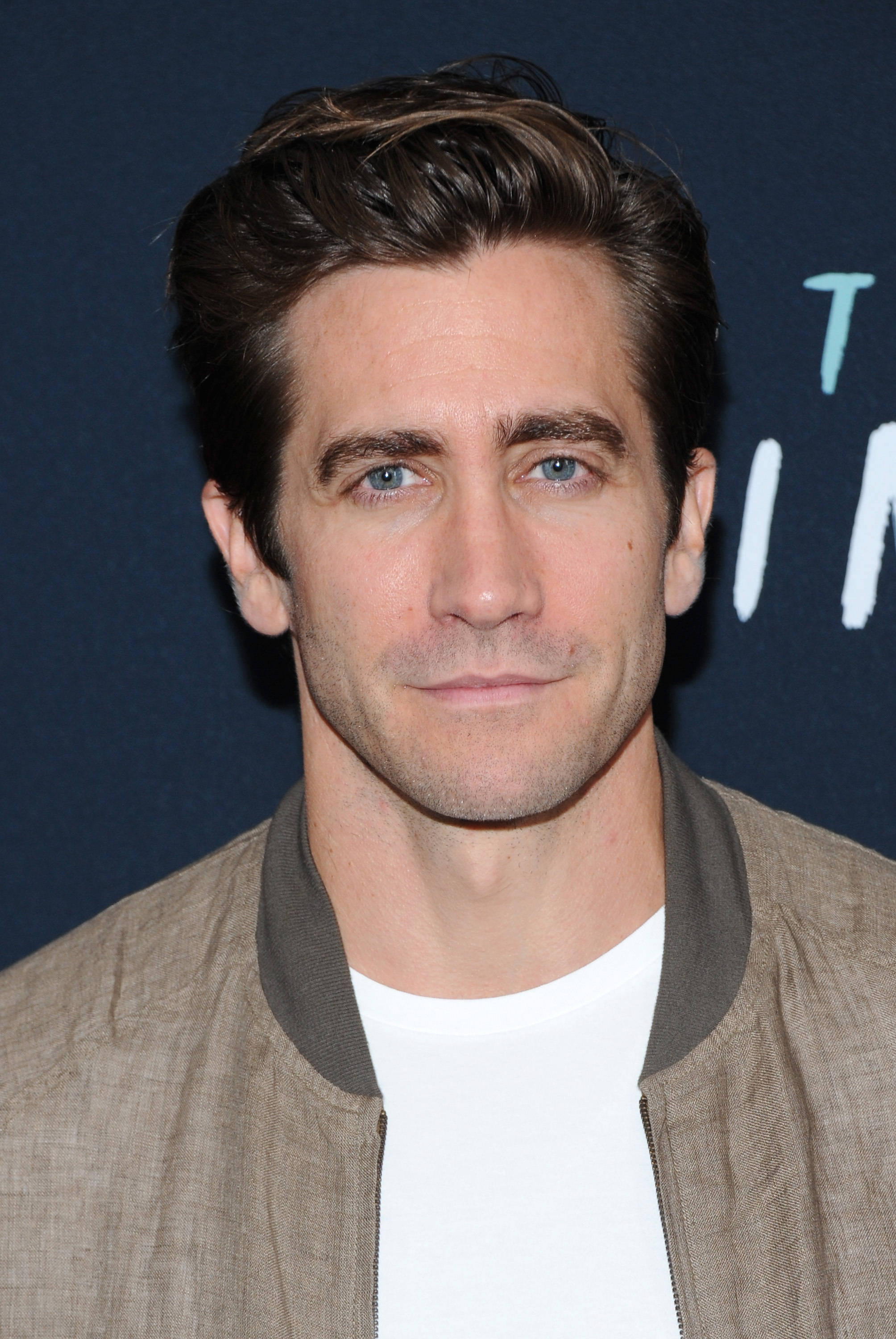jake 桌布 壁紙 — 演員傑克·葛倫霍的close-up肖像，藍眼睛、時尚髮型，身穿米色bomber外套內搭白色T恤，背景為深藍色。