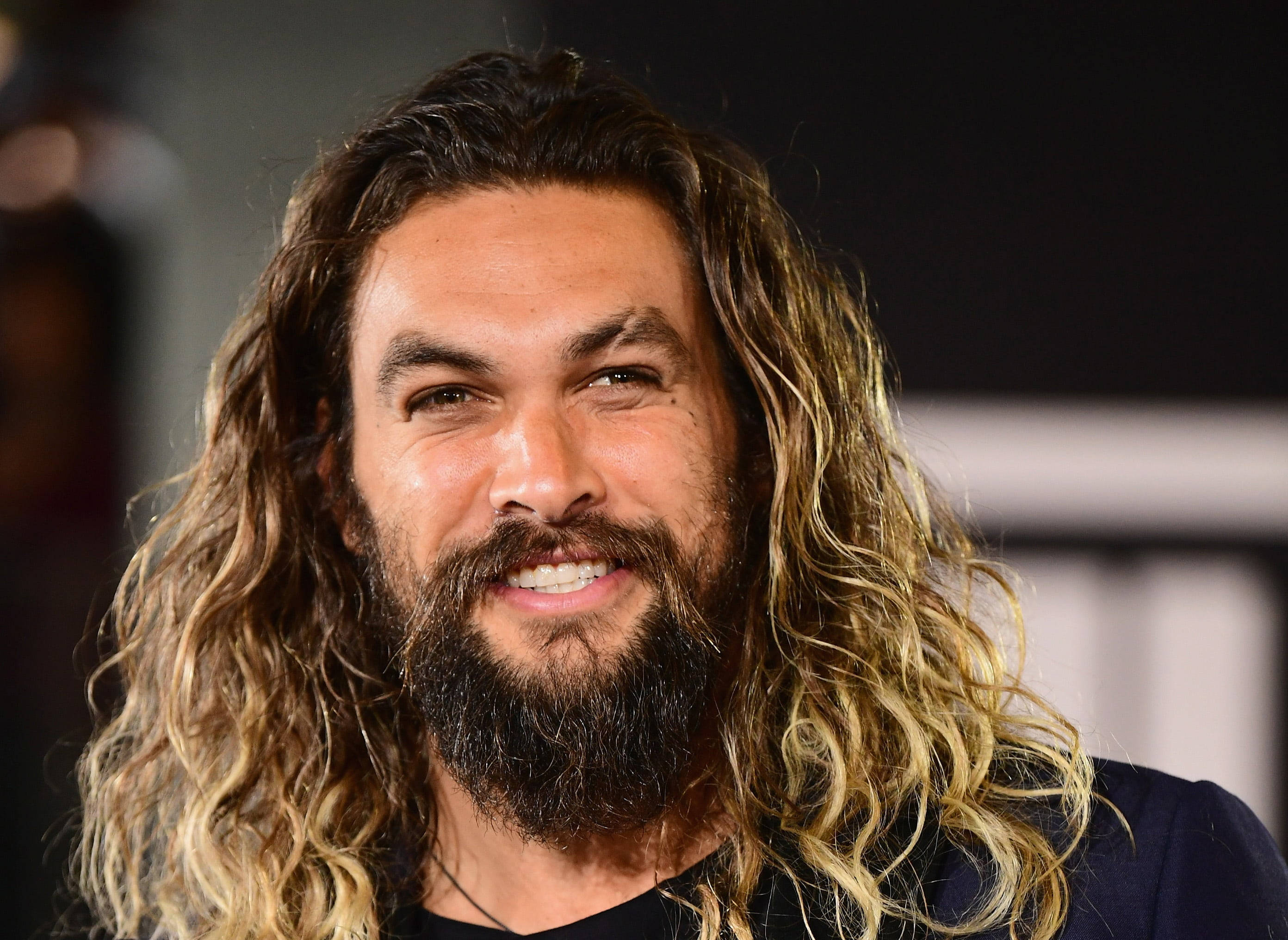 Smile 桌布 壁紙 — 演員傑森·摩莫亞（Jason Momoa）留著長長的金色波浪髮和濃密鬍鬚，在模糊的背景前溫暖地微笑。