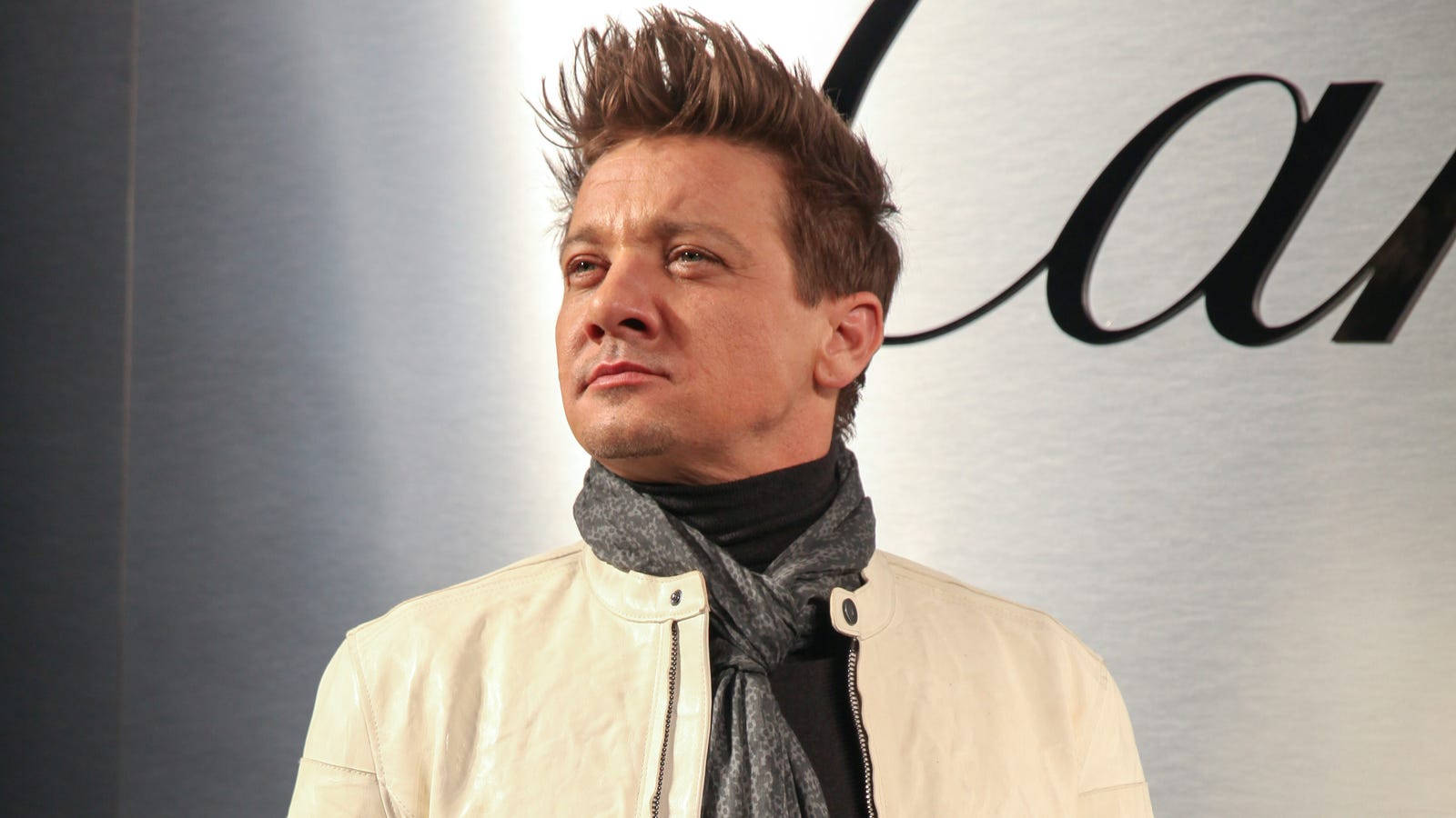 Jeremy Renner 桌布 壁紙 — 演員傑瑞米·雷納身穿米色外套和灰色圍巾，站在帶有黑色標誌的時尚銀色牆壁前。
