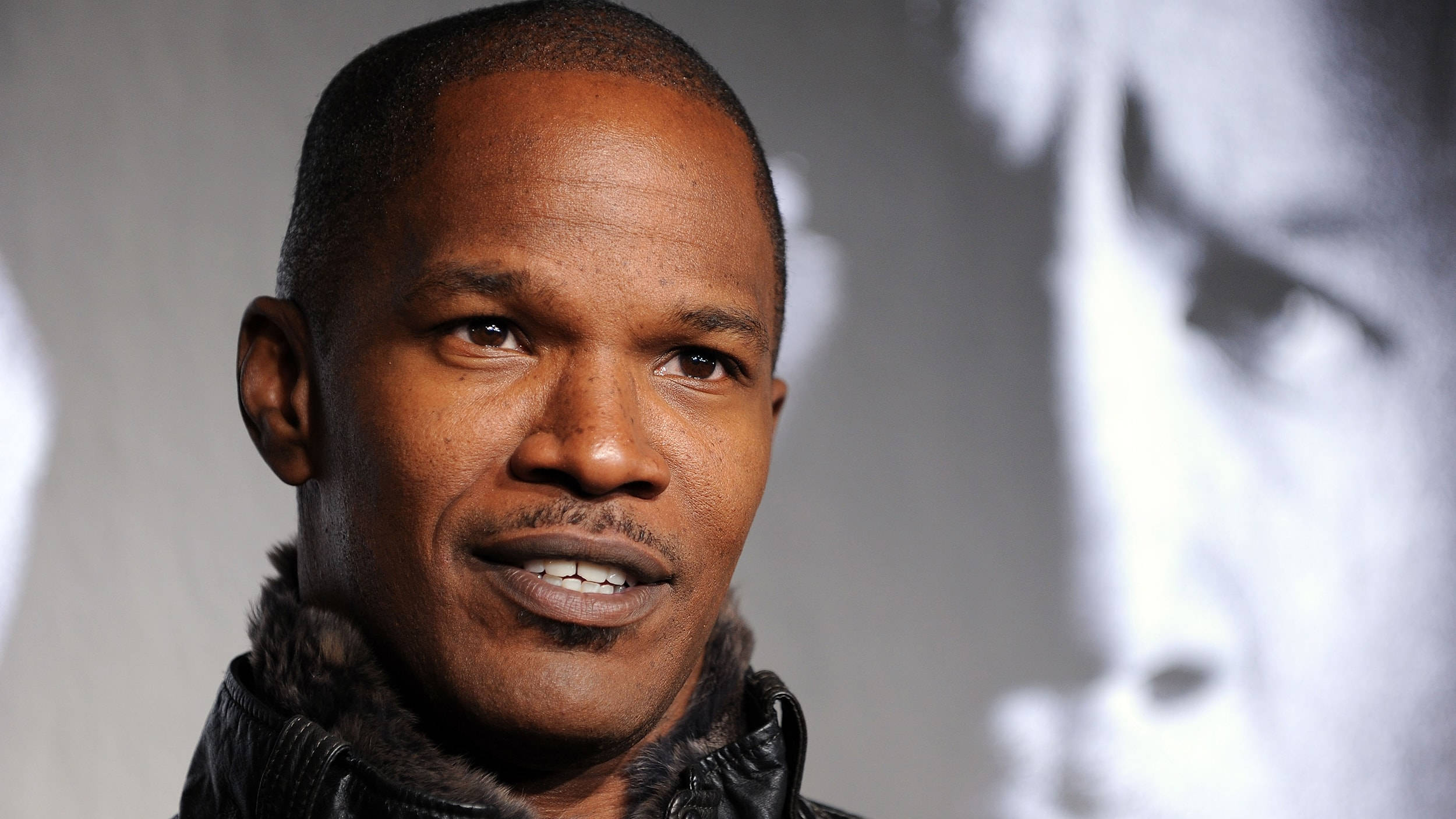 Jamie Foxx 桌布 壁紙 — 演員傑米·福克斯的特寫肖像，穿著帶毛領的深色外套，在模糊的灰階背景前微微微笑。