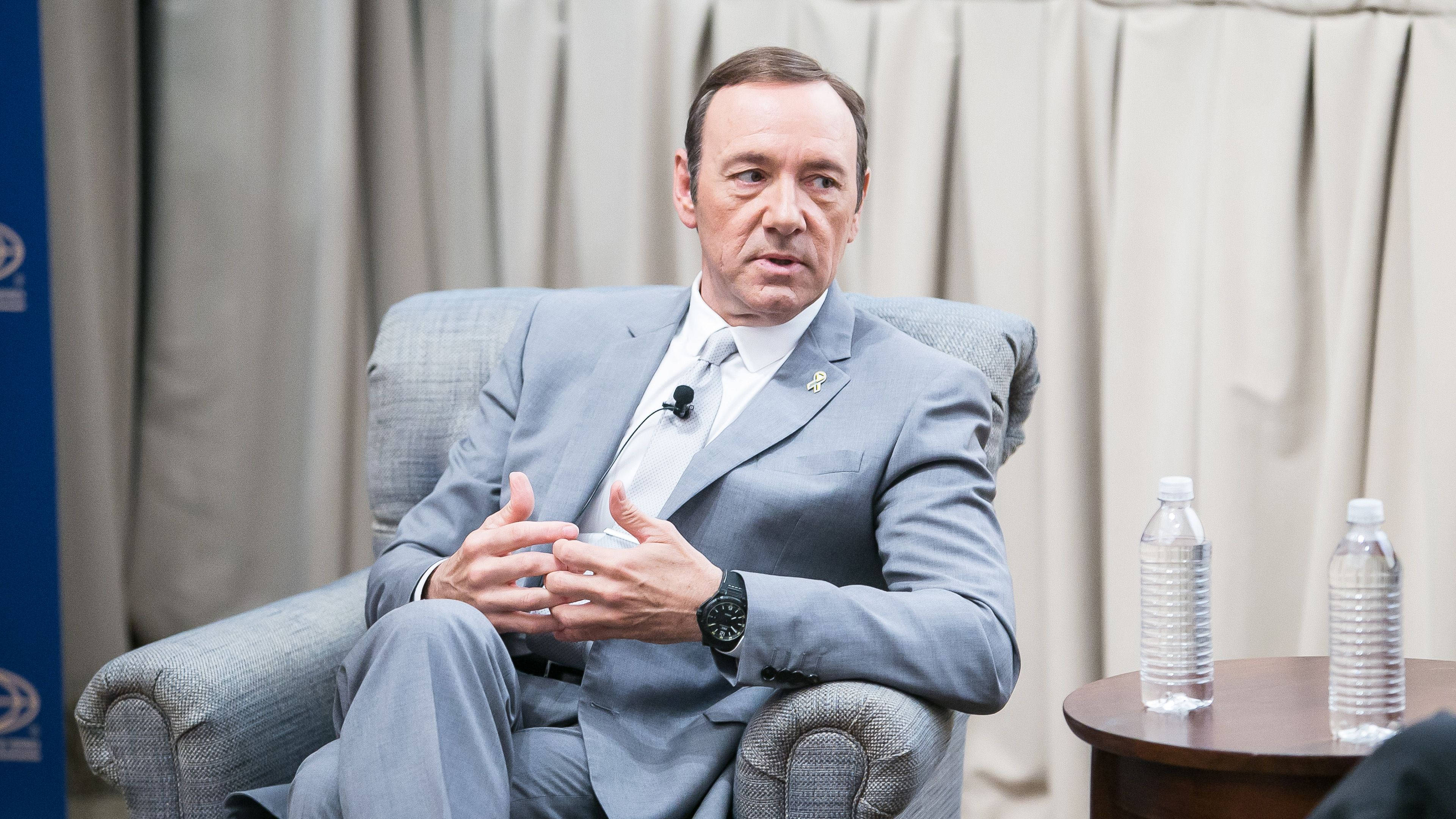 actor 桌布 壁紙 — 演員凱文史貝西坐在灰色扶手椅上，身穿淺灰色西裝，在一個活動中說話時做手勢，旁邊的桌子上有水瓶。