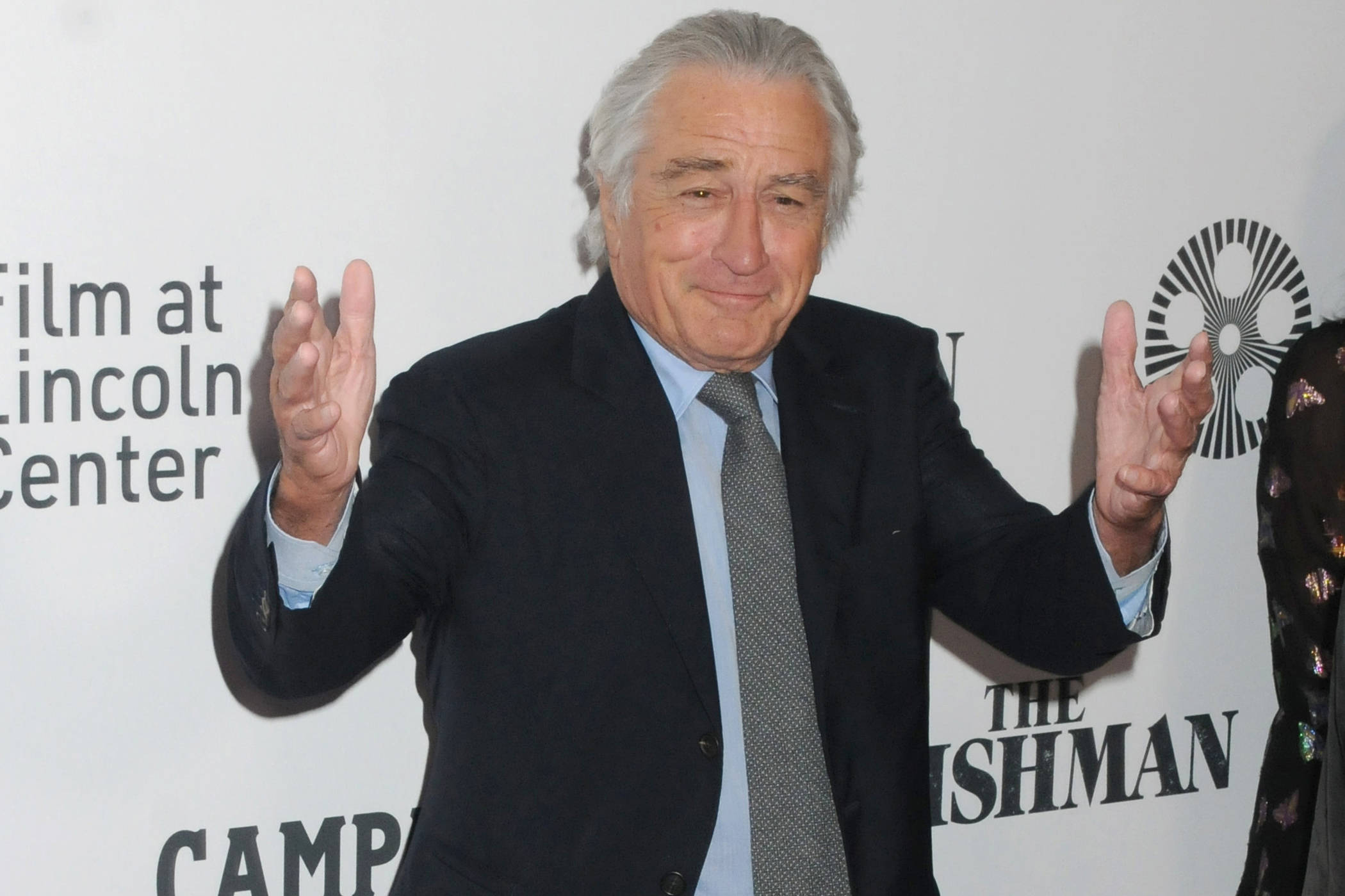 Robert De Niro 桌布 壁紙 — 演員勞勃狄尼洛在活動中穿著深色西裝和圖案領帶，雙手舉起做出友善手勢，溫暖地微笑。