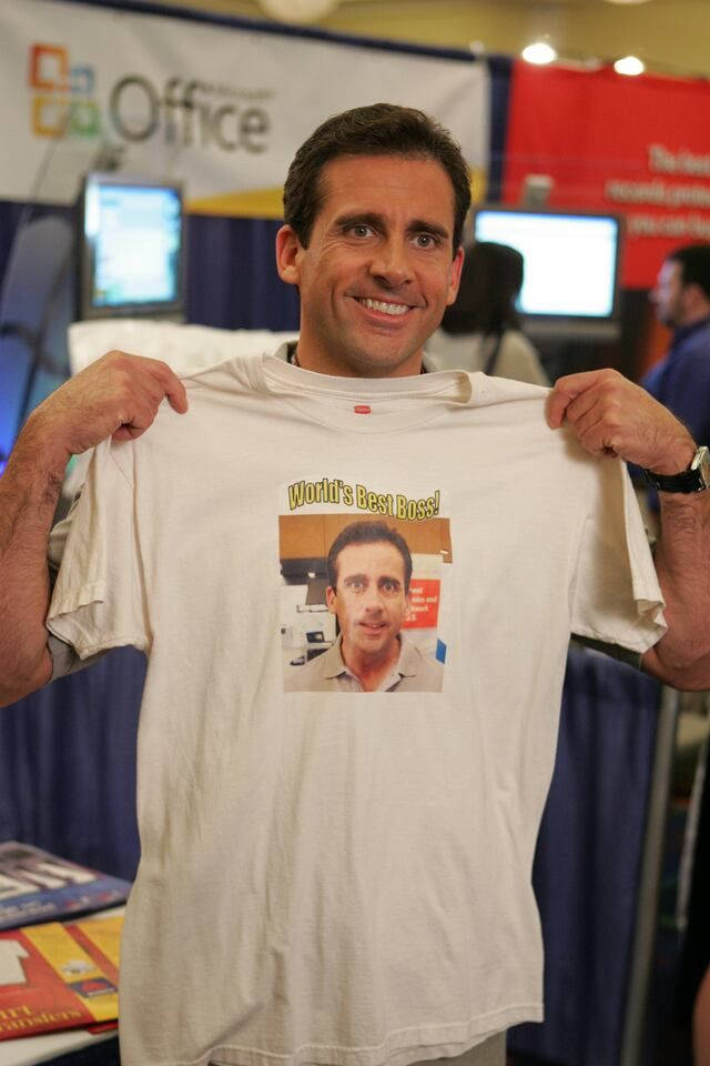 Michael Scott 桌布 壁紙 — 演員史提夫·卡瑞爾在會議攤位微笑著舉起一件印有自己照片和文字「World's Best Boss」的白色T恤。