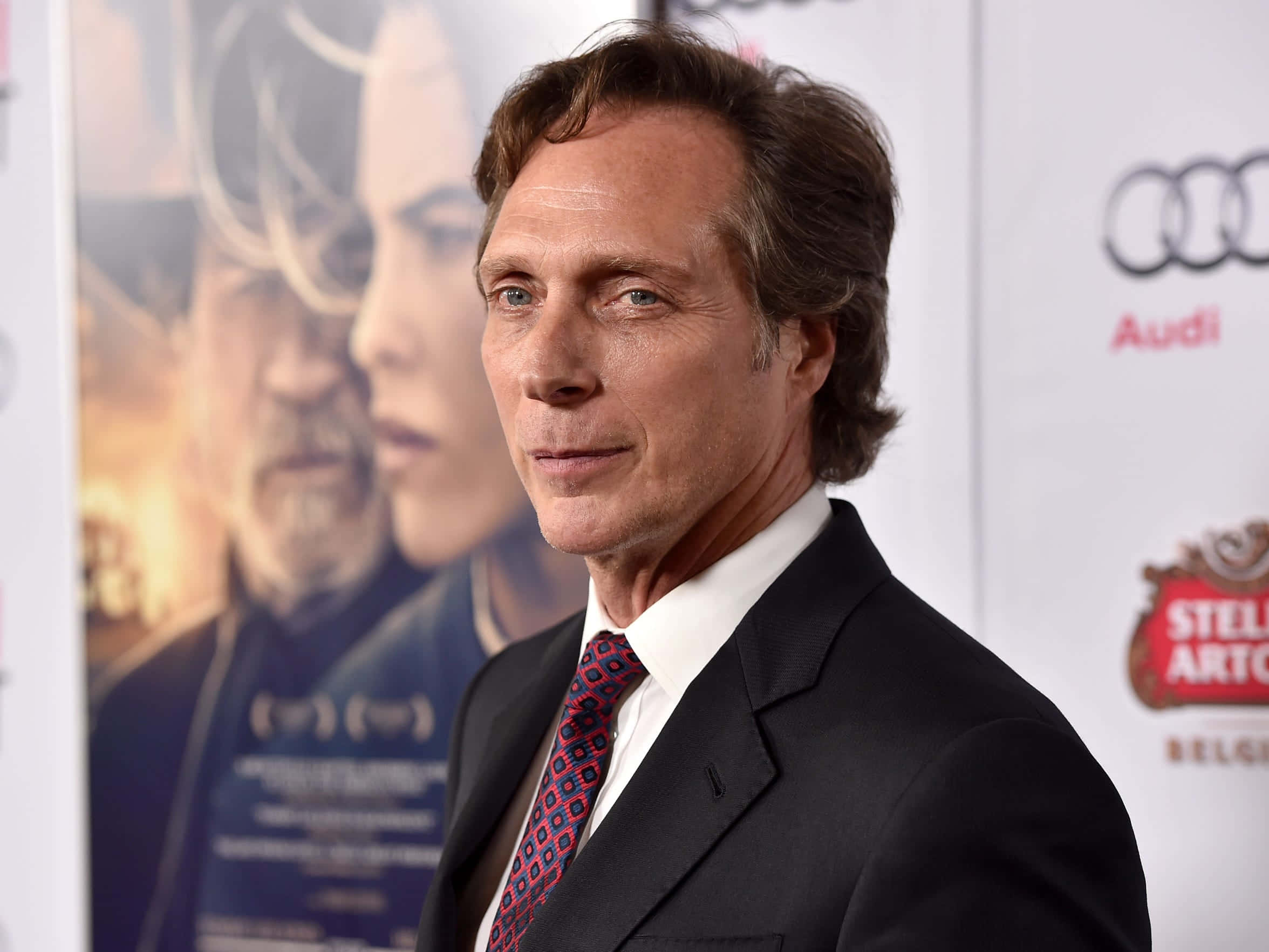 William Fichtner 桌布 壁紙 — 演員威廉·菲希特納身穿俐落黑色西裝和圖案領帶，在紅地毯活動中擺姿勢，身後有電影海報和贊助商標誌。