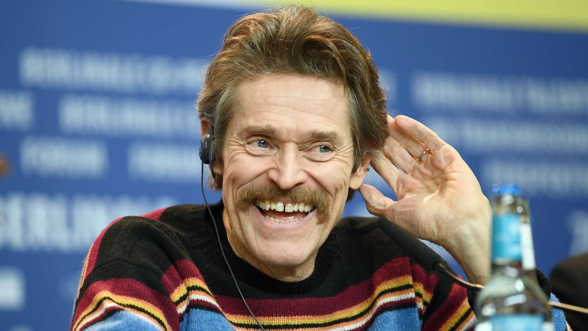 Willem Dafoe 桌布 壁紙 — 演員威廉·達佛在記者會上戴著耳機，身穿彩色條紋毛衣，露出燦爛的笑容。