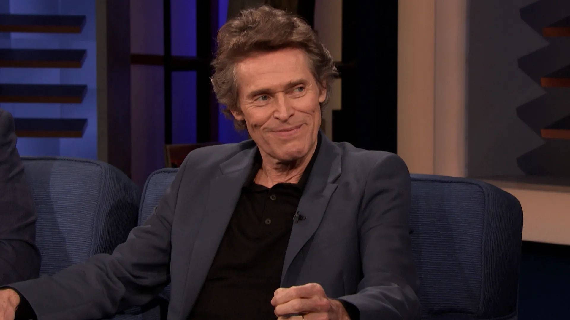 Willem Dafoe 桌布 壁紙 — 演員威廉·達佛在訪談期間放鬆地坐在藍色攝影棚椅子上，帶著一絲微笑。