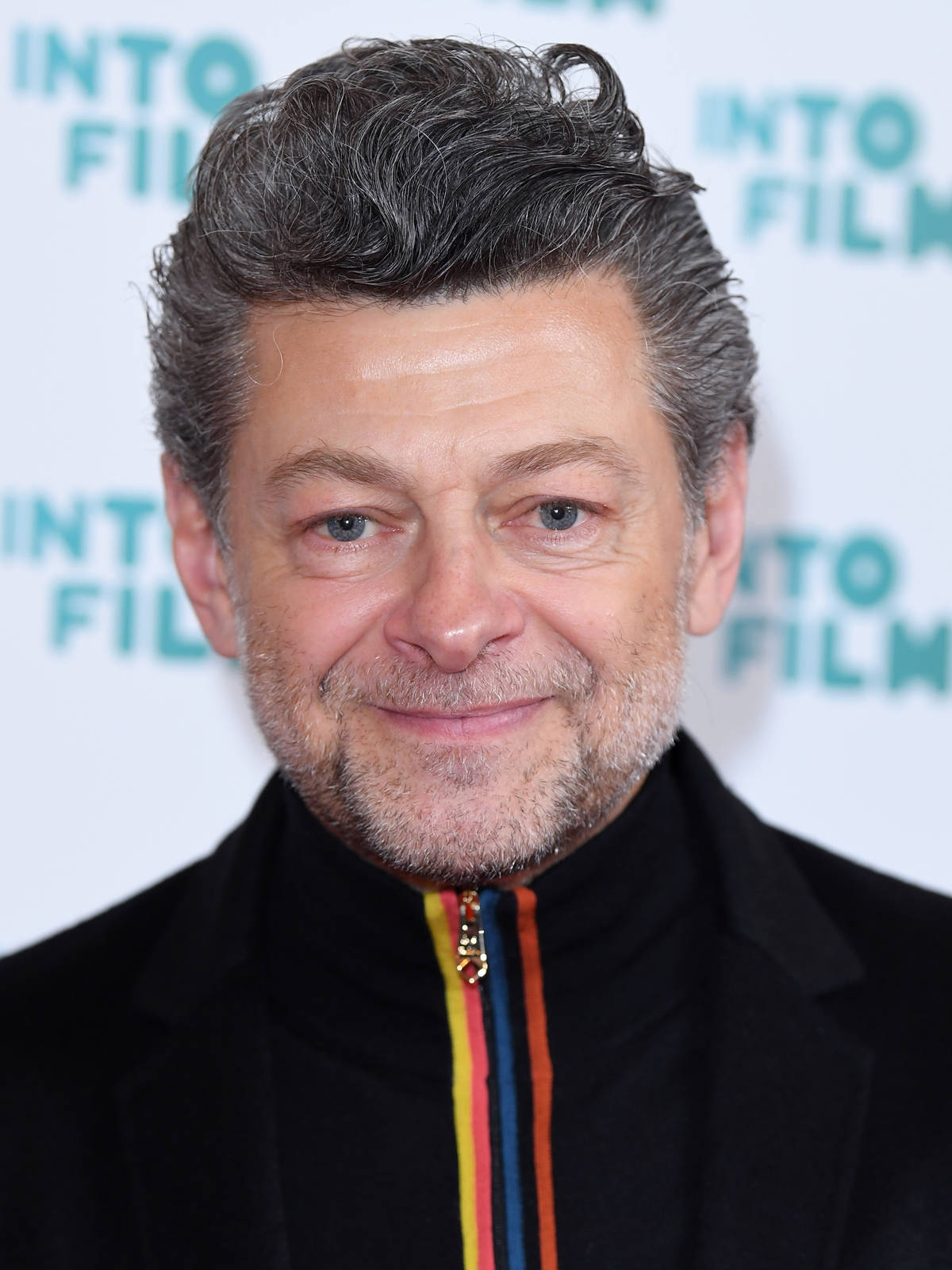 actor 桌布 壁紙 — 演員安迪·瑟克斯（Andy Serkis）穿著帶有彩色拉鏈的黑色外套，在白色背景配上青綠色文字前，對著鏡頭溫暖微笑。