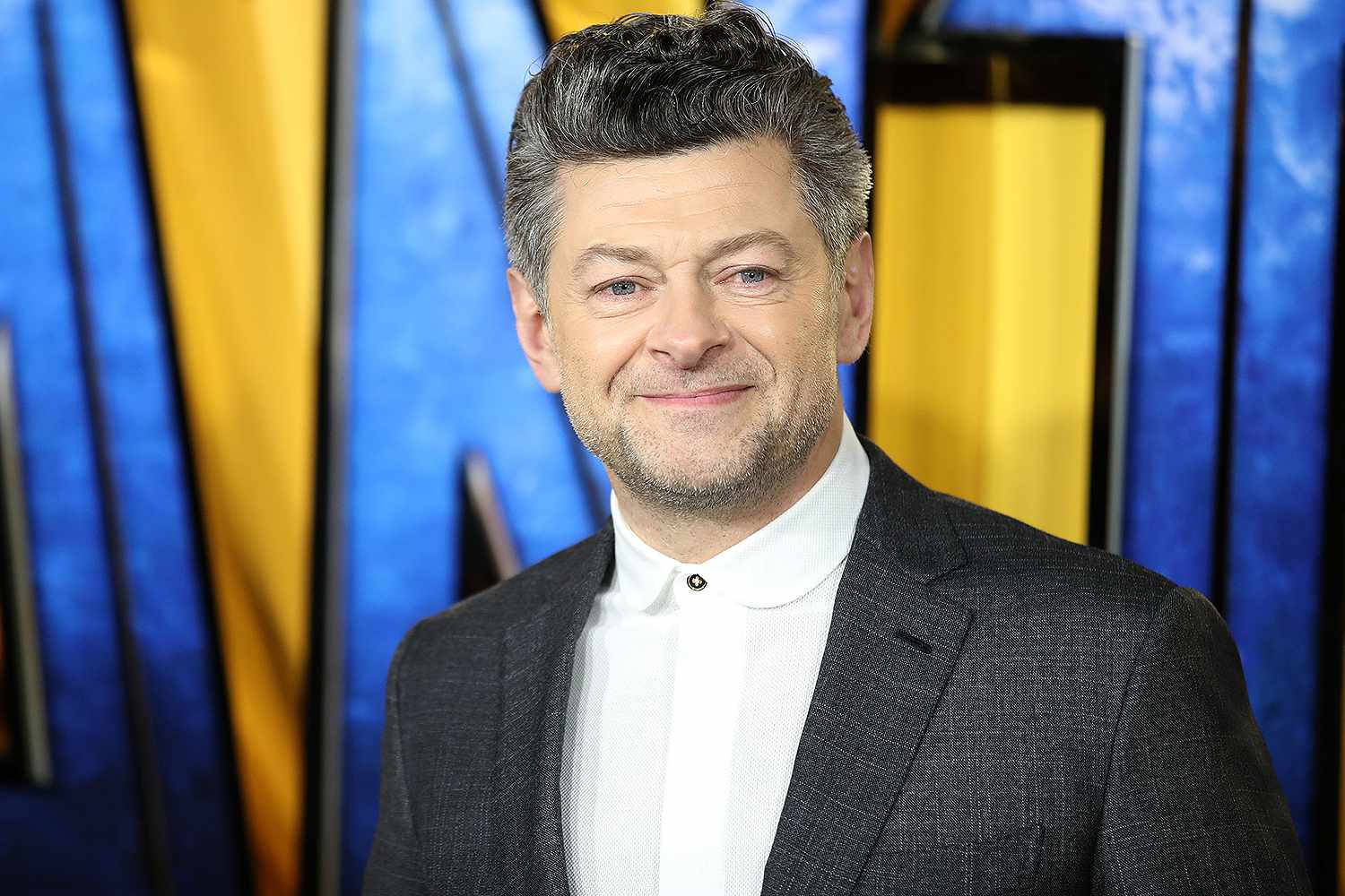 actor 桌布 壁紙 — 演員安迪·瑟克斯在充滿活力的藍黃條紋背景下，身穿深色西裝和白色襯衫，溫暖地微笑。