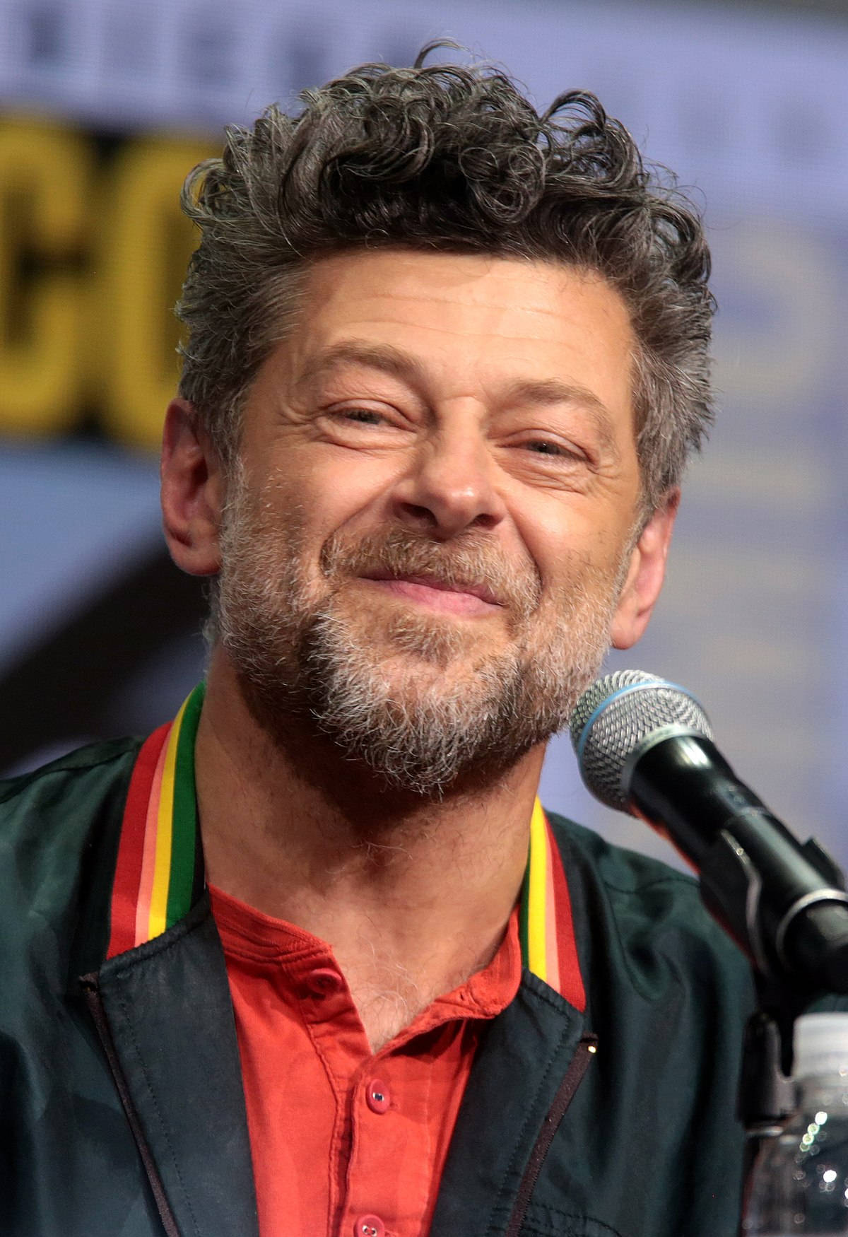 actor 桌布 壁紙 — 演員安迪·瑟克斯在座談會上溫暖地微笑，面前有麥克風，身穿紅色襯衫和綠色外套。