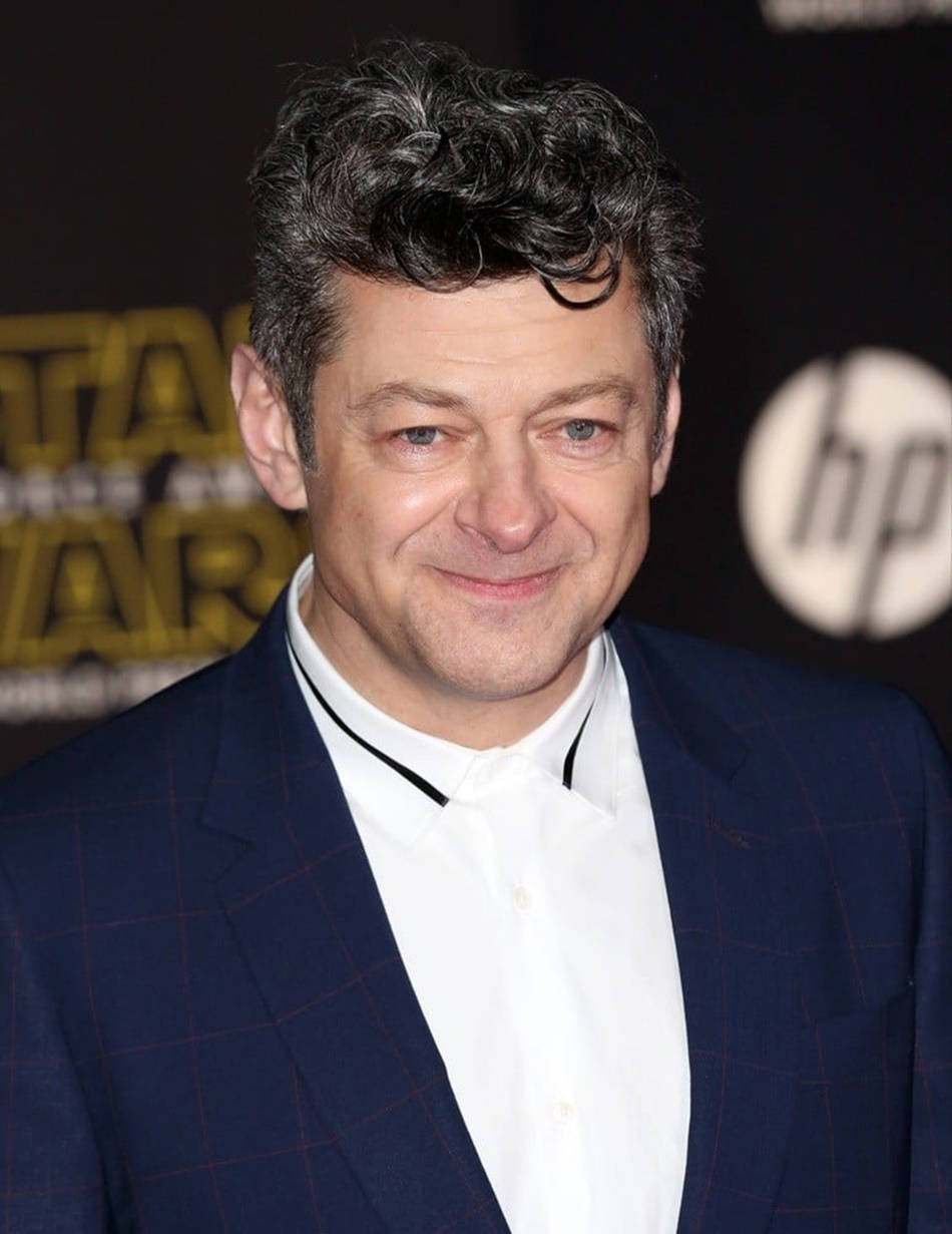 actor 桌布 壁紙 — 演員安迪·瑟克斯在星際大戰活動中，身後有HP品牌標誌，身穿海軍藍格紋西裝溫暖微笑。