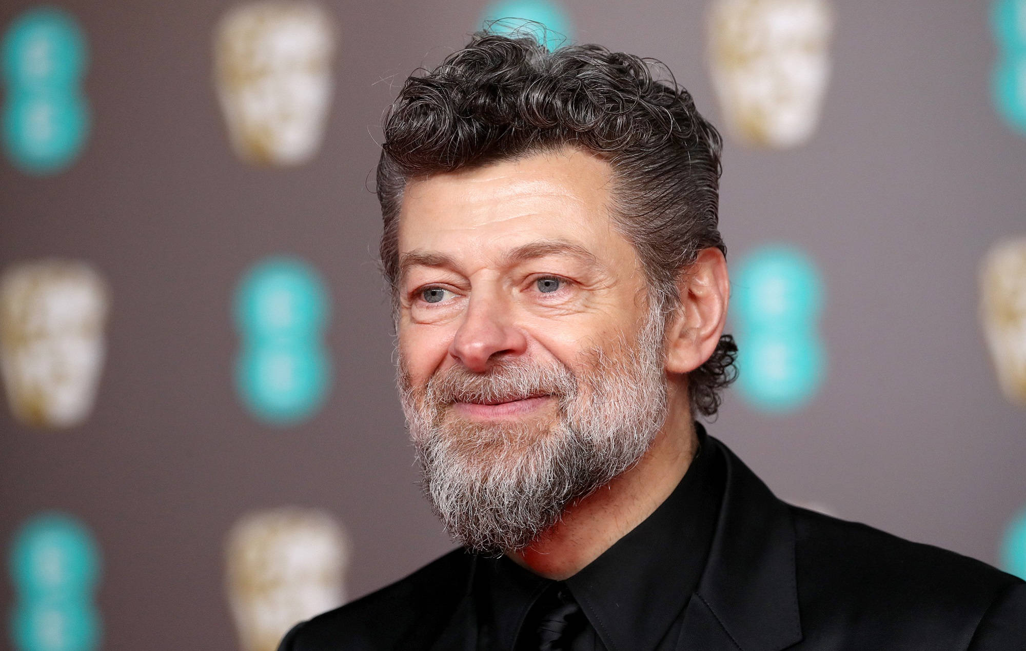 actor 桌布 壁紙 — 演員安迪·瑟克斯穿著黑色西裝，在重複的BAFTA面具背景下溫暖地微笑。