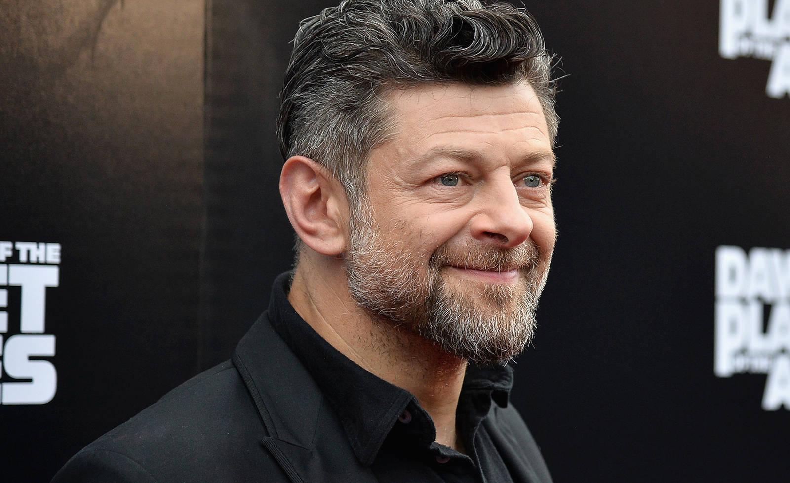 actor 桌布 壁紙 — 演員安迪·瑟克斯穿著黑色西裝，在帶有白色文字的深色宣傳背景前溫暖地微笑。