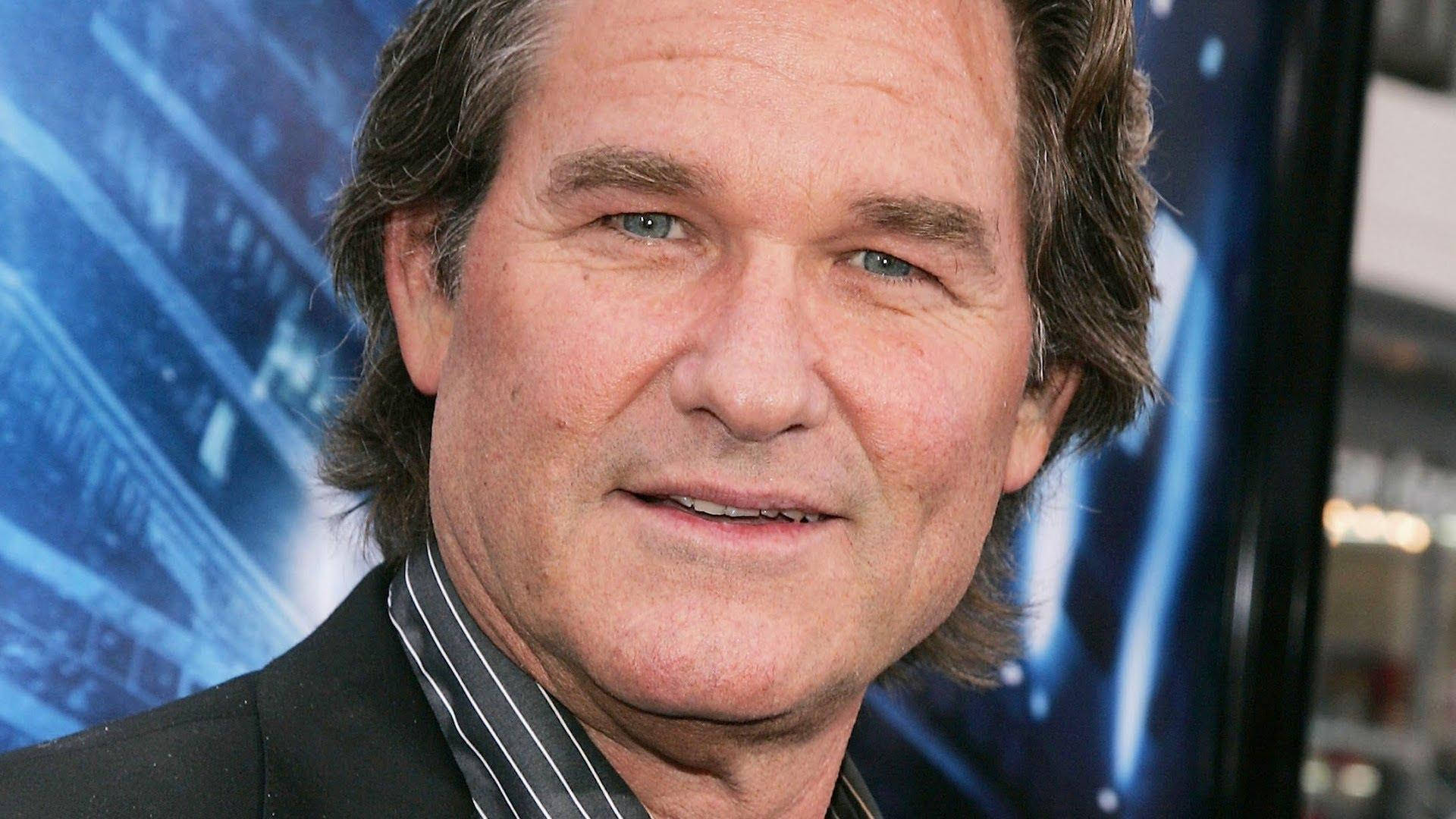 電影動圖 桌布 壁紙 — 演員寇特·羅素（Kurt Russell）身穿黑色西裝，在藍色電影海報背景下溫暖微笑的特寫肖像。