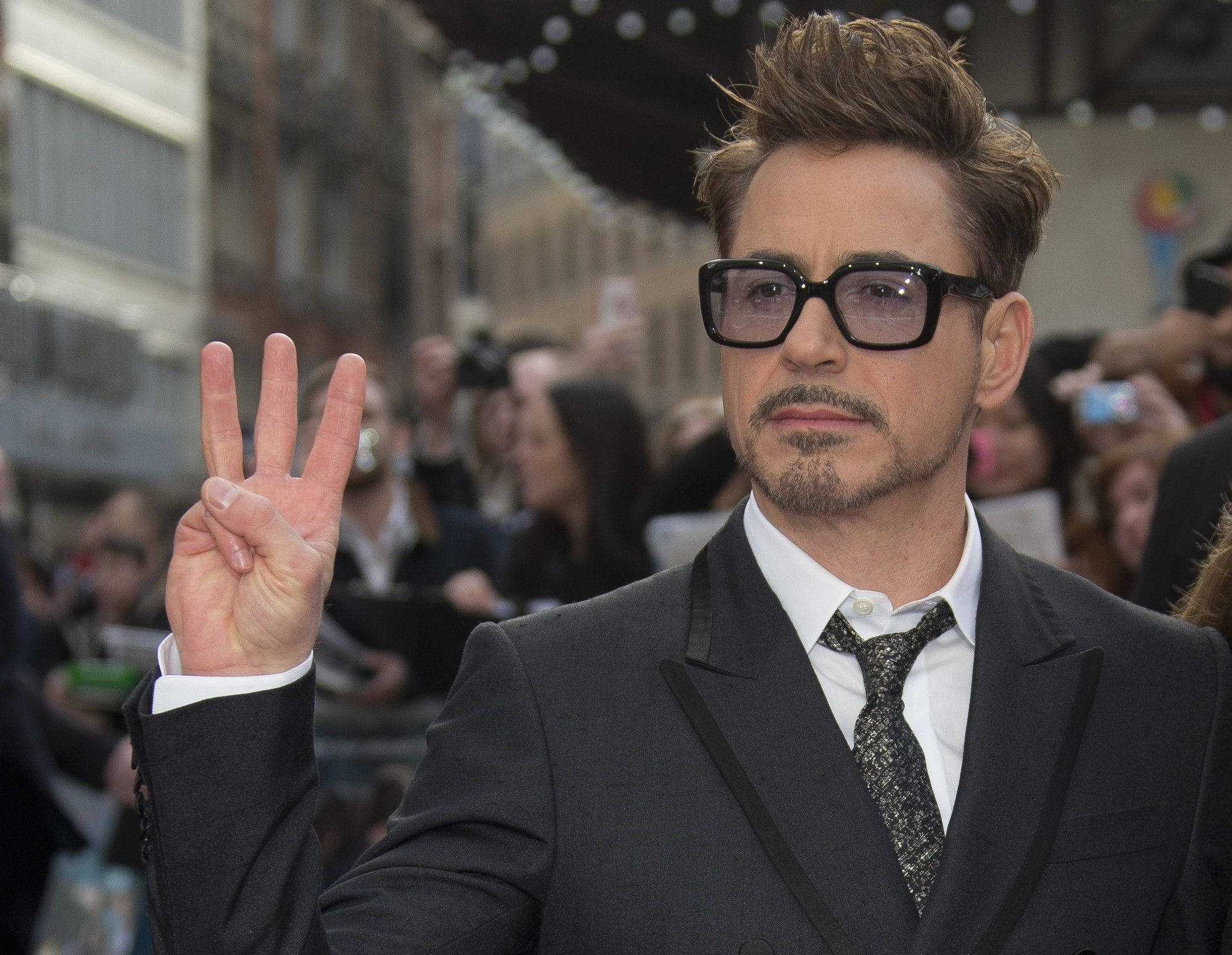 小勞勃·道尼 桌布 壁紙 — 演員小勞勃·道尼（Robert Downey Jr.）身穿筆挺西裝和眼鏡，在一個熙熙攘攘的戶外活動中向粉絲和鏡頭比出和平手勢。