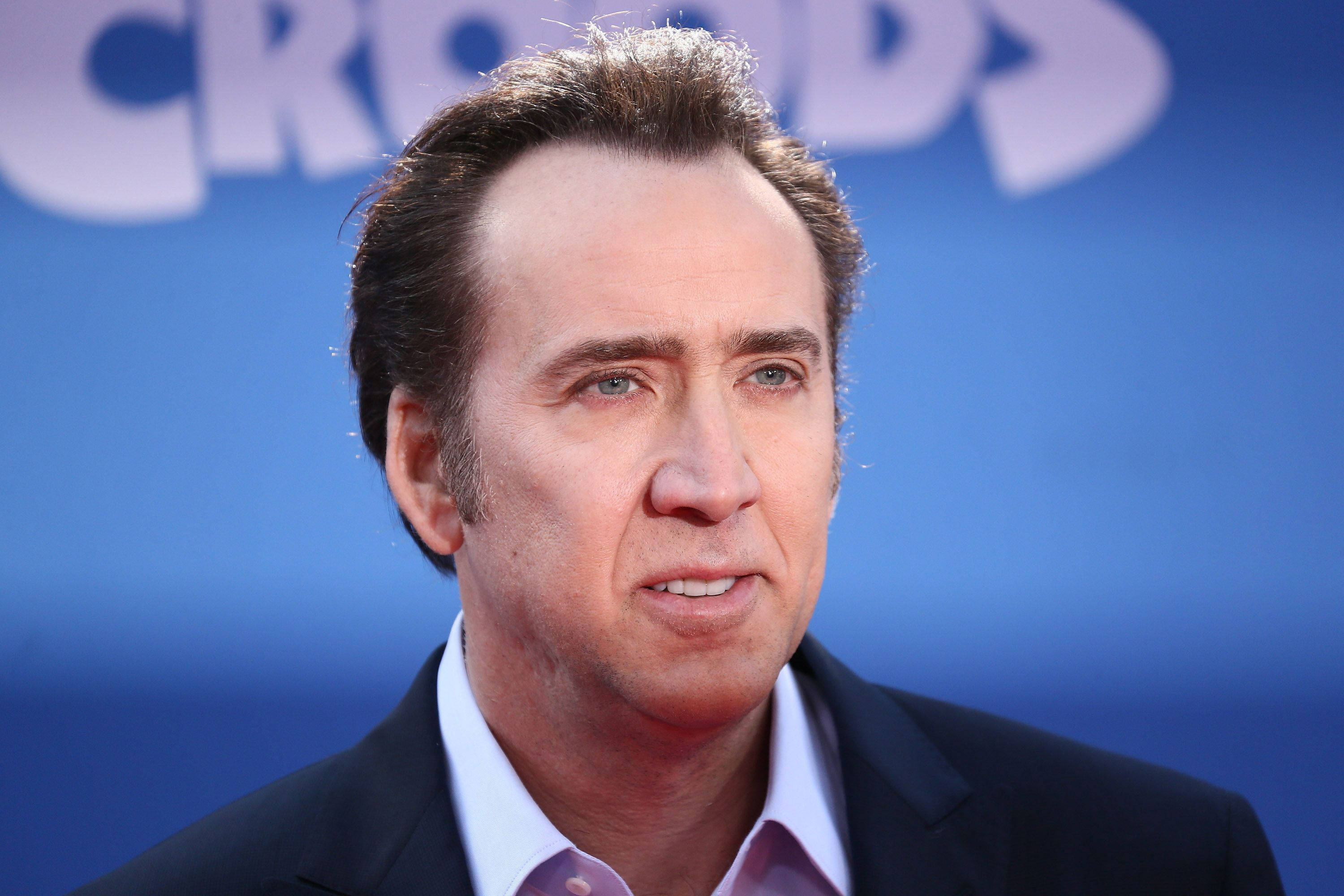 Nicolas Cage 桌布 壁紙 — 演員尼可拉斯·凱吉身穿深色西裝和淺色襯衫，背景為模糊的藍色，帶有白色字樣。