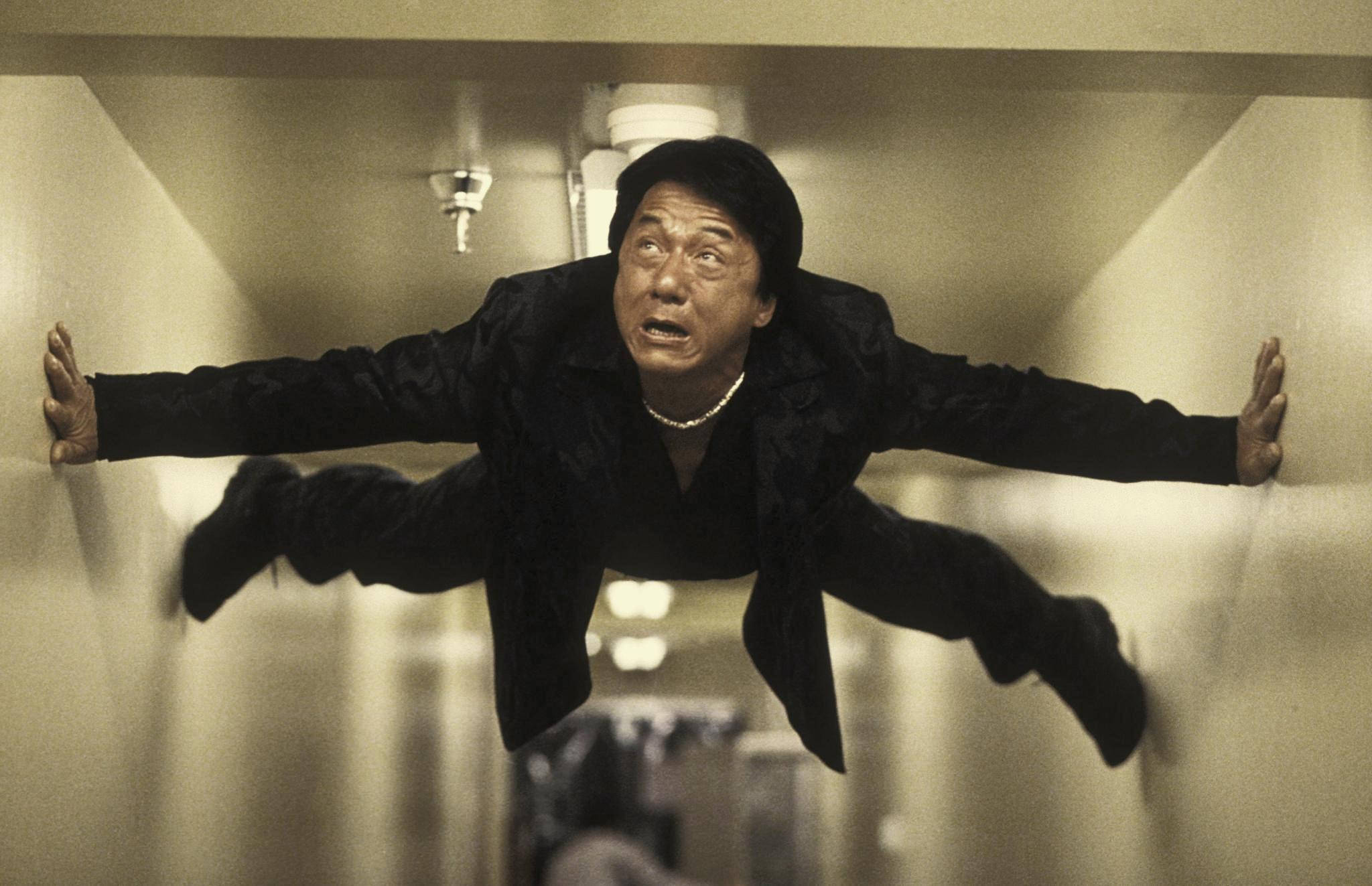 jackie chan 桌布 壁紙 — 演員成龍在走廊中懸浮半空，手腳張開，身穿黑色西裝和珍珠項鍊。