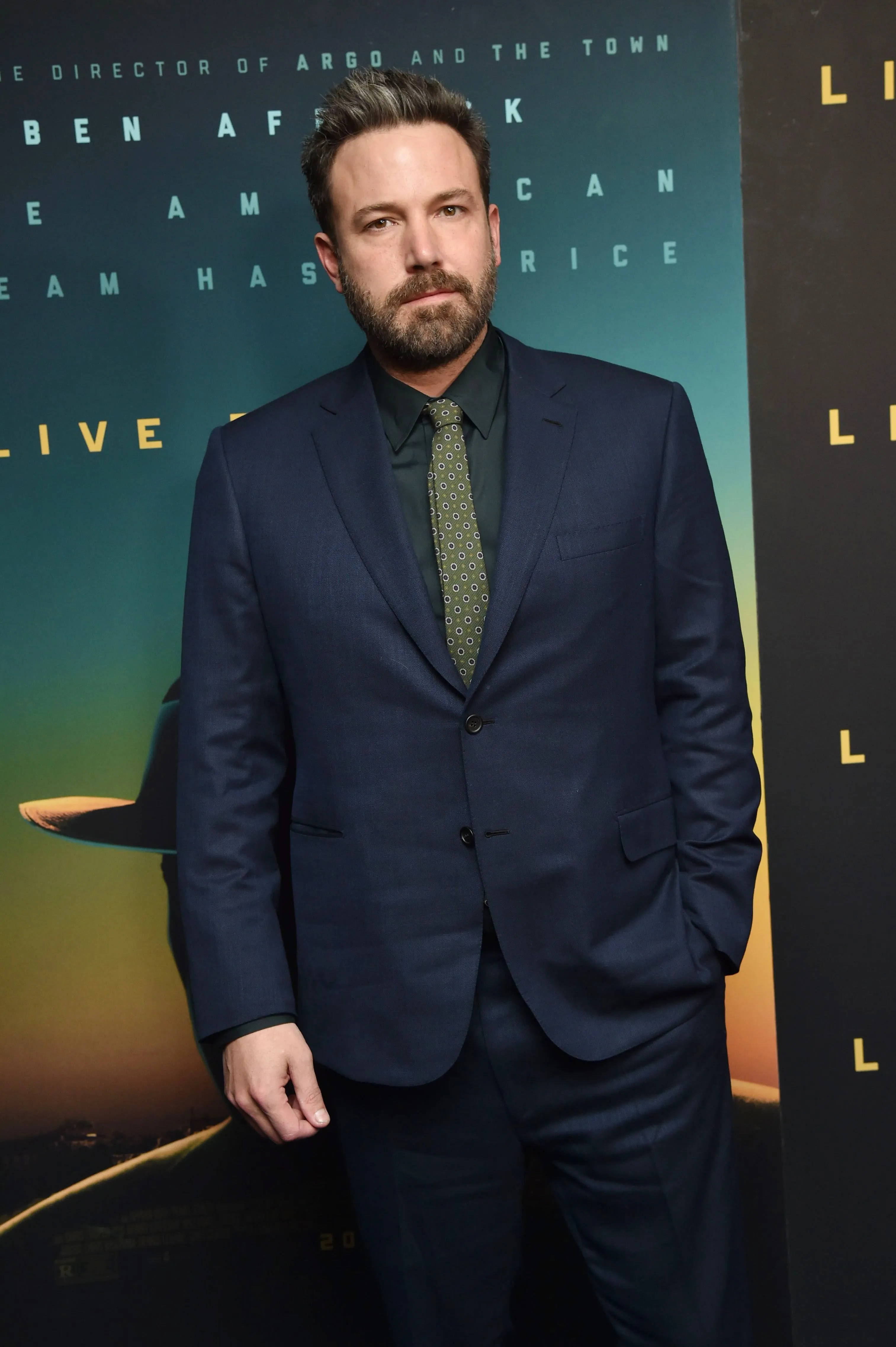 Ben Affleck 桌布 壁紙 — 演員班·艾佛列克自信地穿著海軍藍西裝，站在電影首映背景前，背景有發光文字和溫暖漸變燈光。