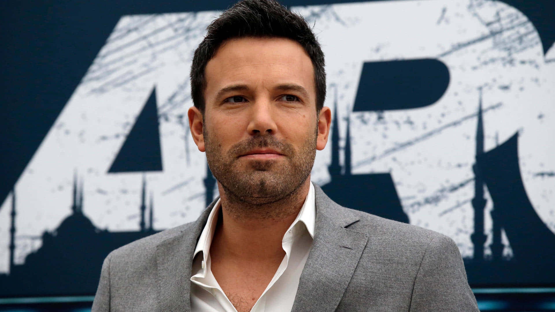Ben Affleck 桌布 壁紙 — 演員班·艾佛列克身穿灰色西裝和白色襯衫，站在風格化的電影海報背景前。