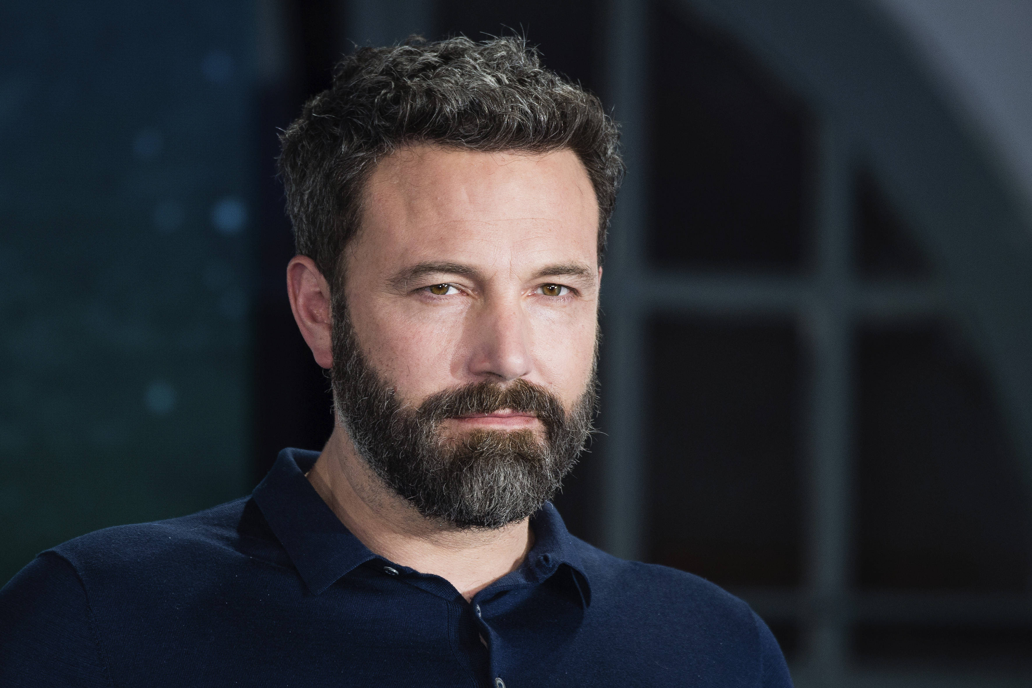 Ben Affleck 桌布 壁紙 — 演員班艾佛列克的特寫肖像，表情嚴肅，身穿深藍色Polo衫，背景模糊昏暗。