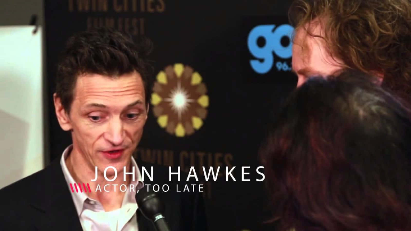 John Hawkes 桌布 壁紙 — 演員約翰·霍克斯在一個帶有標誌的深色背景的電影節新聞發布會上對著麥克風講話。