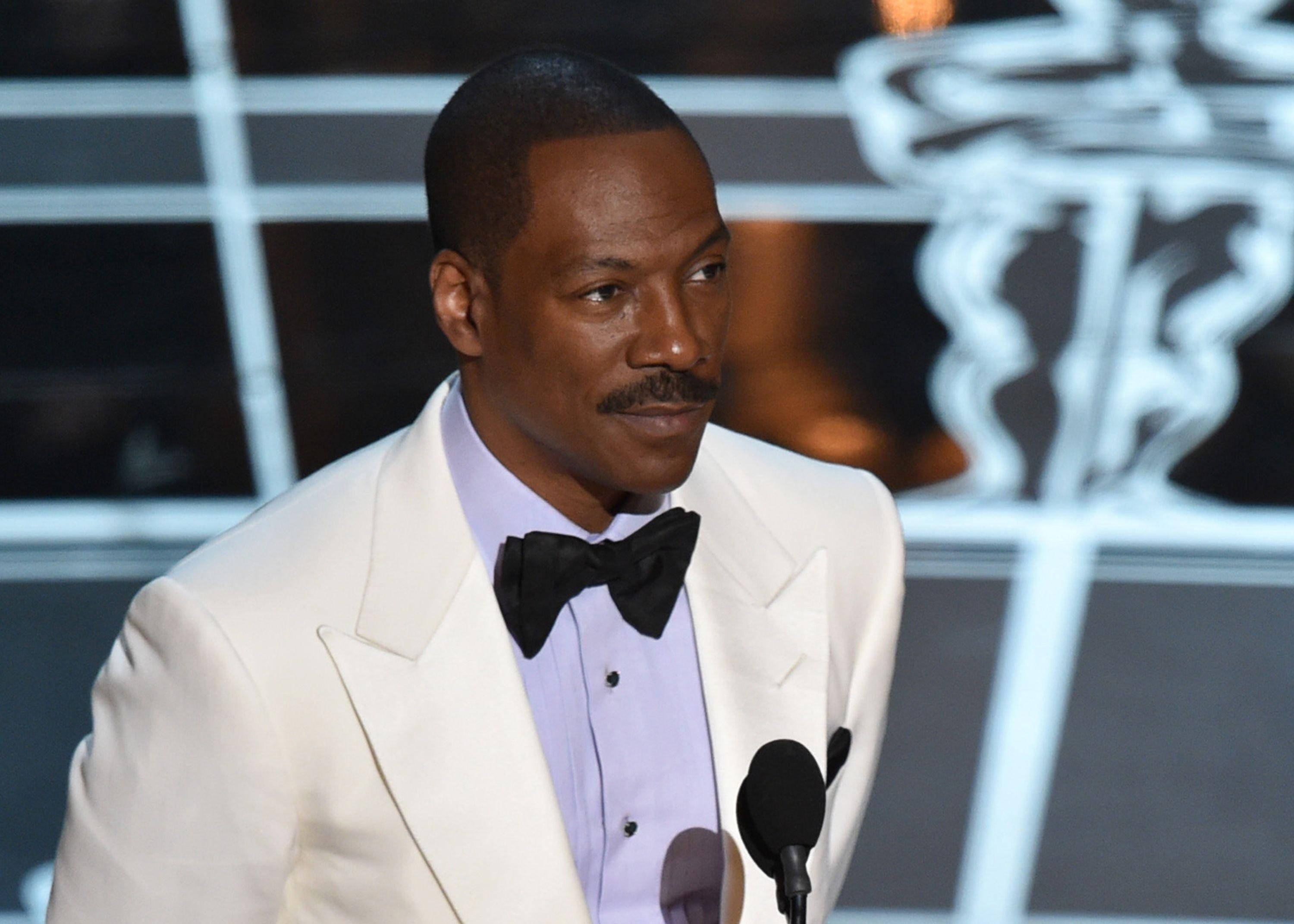 eddie murphy 桌布 壁紙 — 演員艾迪·墨菲身穿白色燕尾服配黑色領結，站在以奧斯卡獎座為背景的舞台講台上。