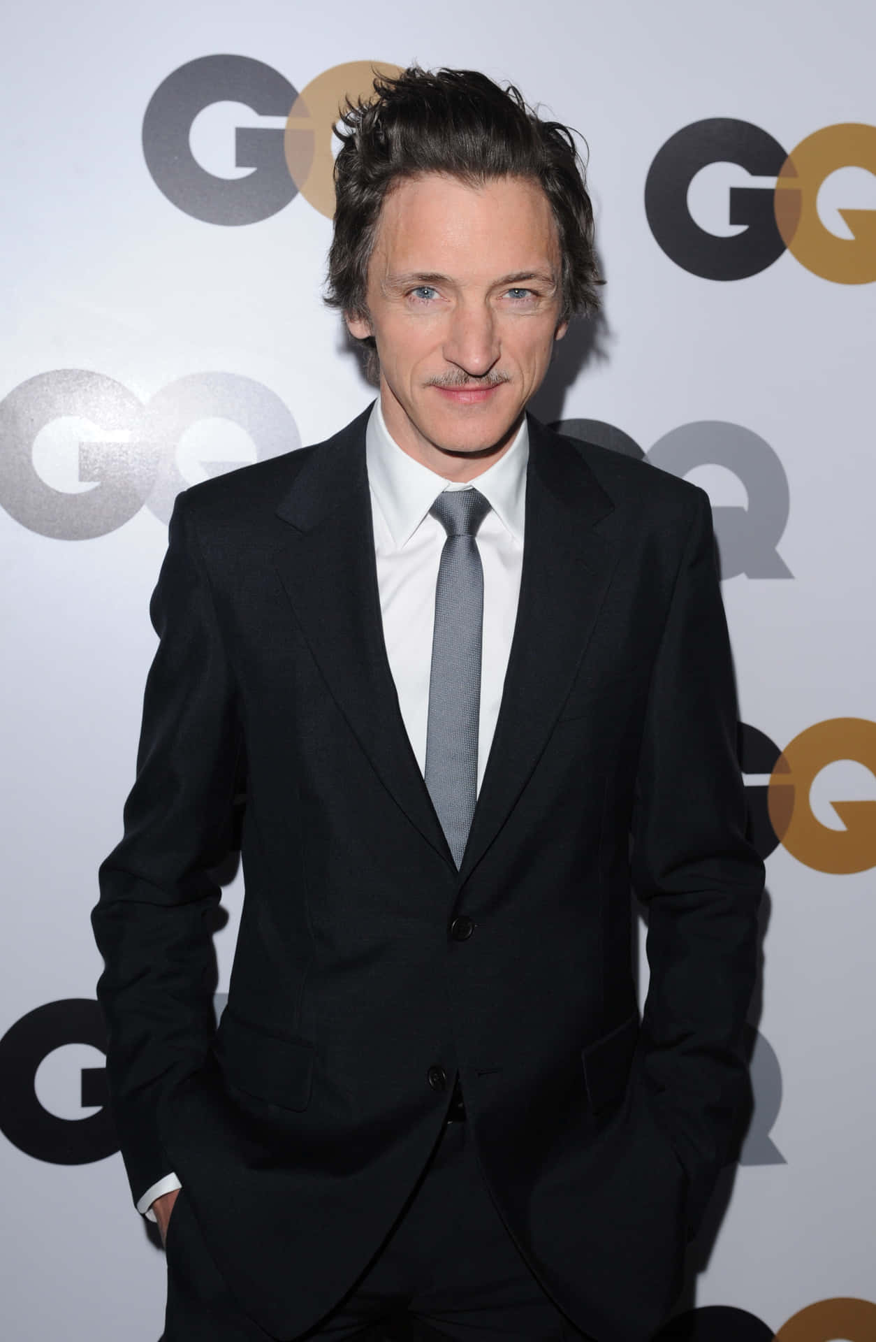 John Hawkes 桌布 壁紙 — 演員身穿俐落黑色西裝和灰色領帶，在活動中自信地以GQ品牌背景擺姿勢。