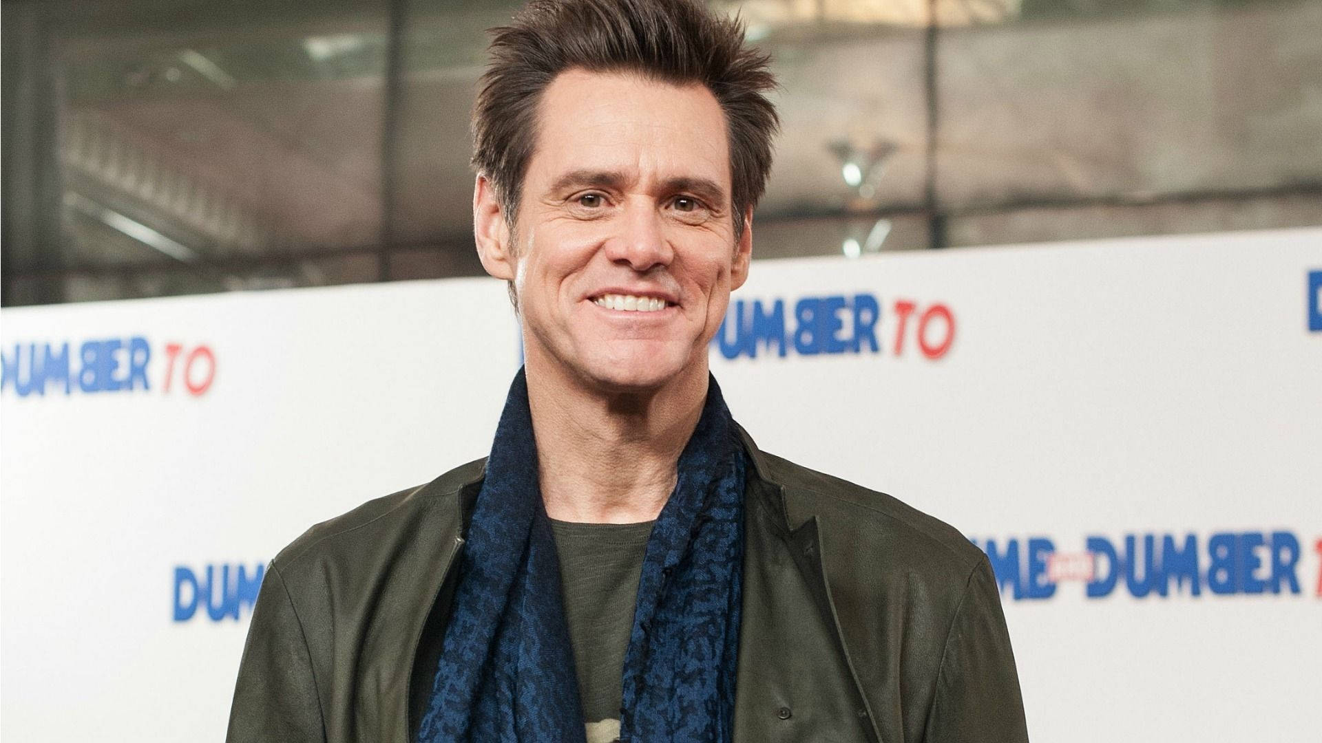 Jim Carrey 桌布 壁紙 — 演員金凱瑞在《阿呆與阿瓜：賤招拆招》首映會上，身穿綠色夾克和藍色圍巾，露出燦爛笑容。
