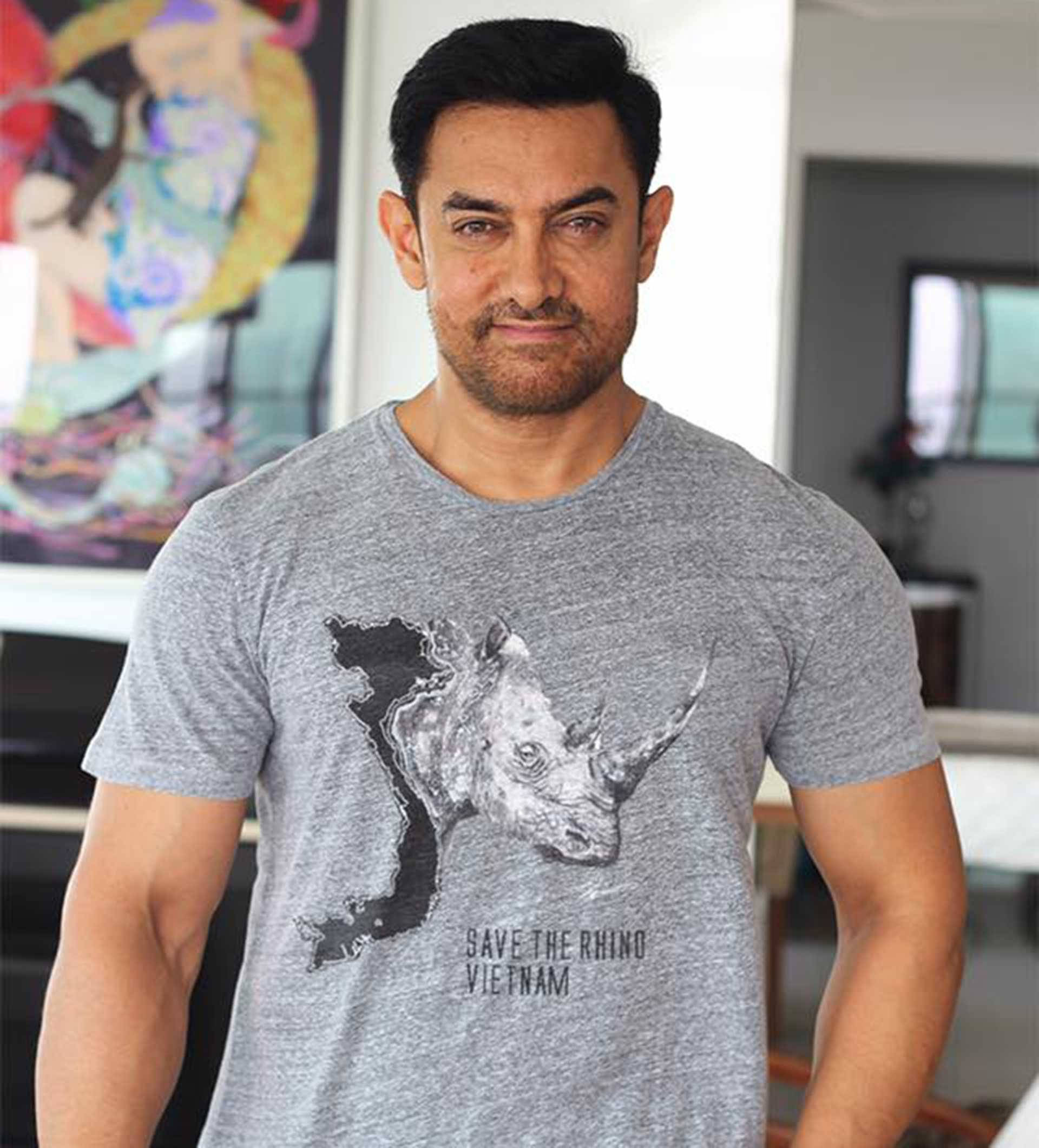 Aamir Khan 桌布 壁紙 — 演員阿米爾·汗自信地站在現代房間裡，身穿灰色T恤，上面有犀牛和文字save the rhino vietnam。