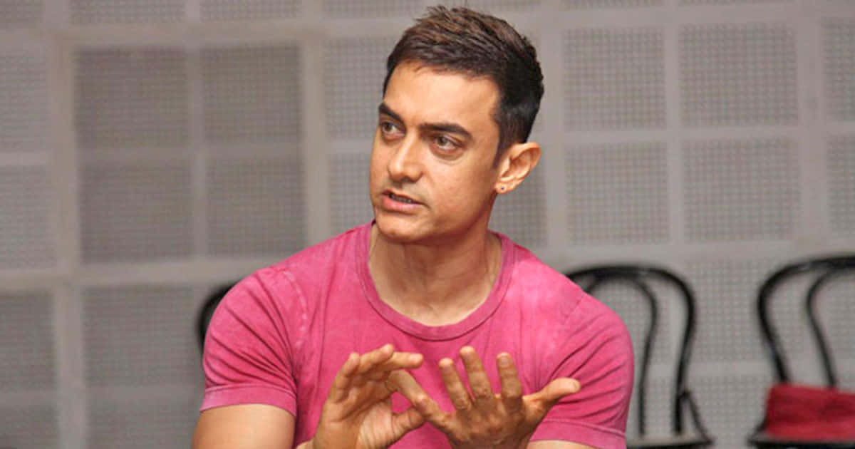 Aamir Khan 桌布 壁紙 — 演員阿米爾汗穿著粉紅色T恤，在網格圖案牆壁前說話並做手勢，身後有椅子。