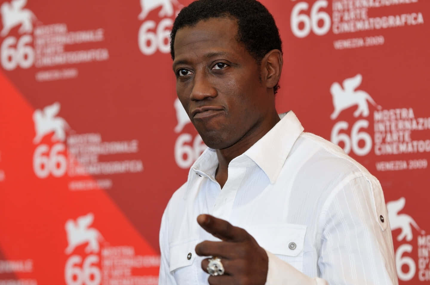 Wesley Snipes 桌布 壁紙 — 演員韋斯利·史奈普斯在第66屆威尼斯影展上自信地擺姿勢，身穿白色襯衫，戴著戒指的手做手勢，背景為紅色品牌背景。