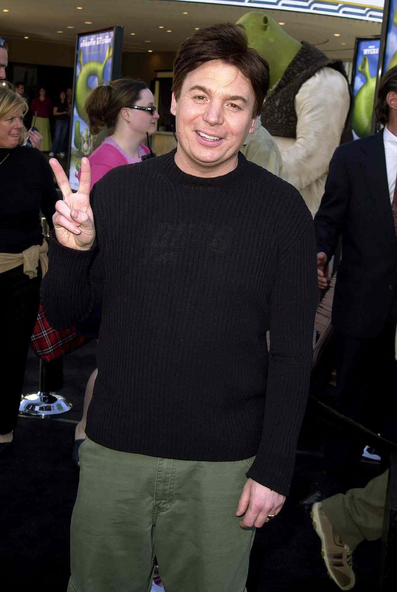 Mike Myers 桌布 壁紙 — 演員麥克邁爾斯微笑並向鏡頭做出和平手勢，身後有史瑞克電影首映的綠色怪物雕像。