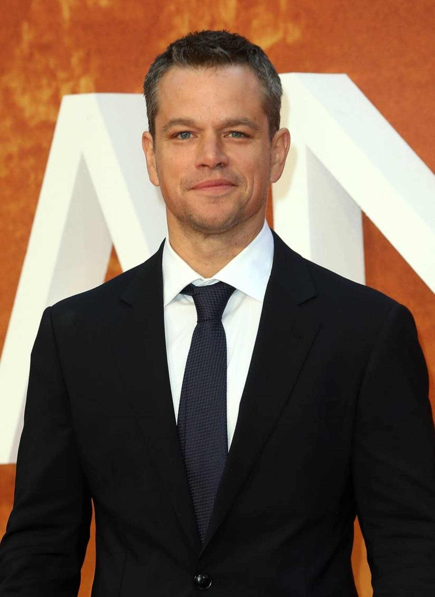 Matt Damon 桌布 壁紙 — 演員麥特戴蒙在溫暖的橙色紋理背景上，自信地穿著俐落的黑色西裝，旁邊有一個大的白色字母。