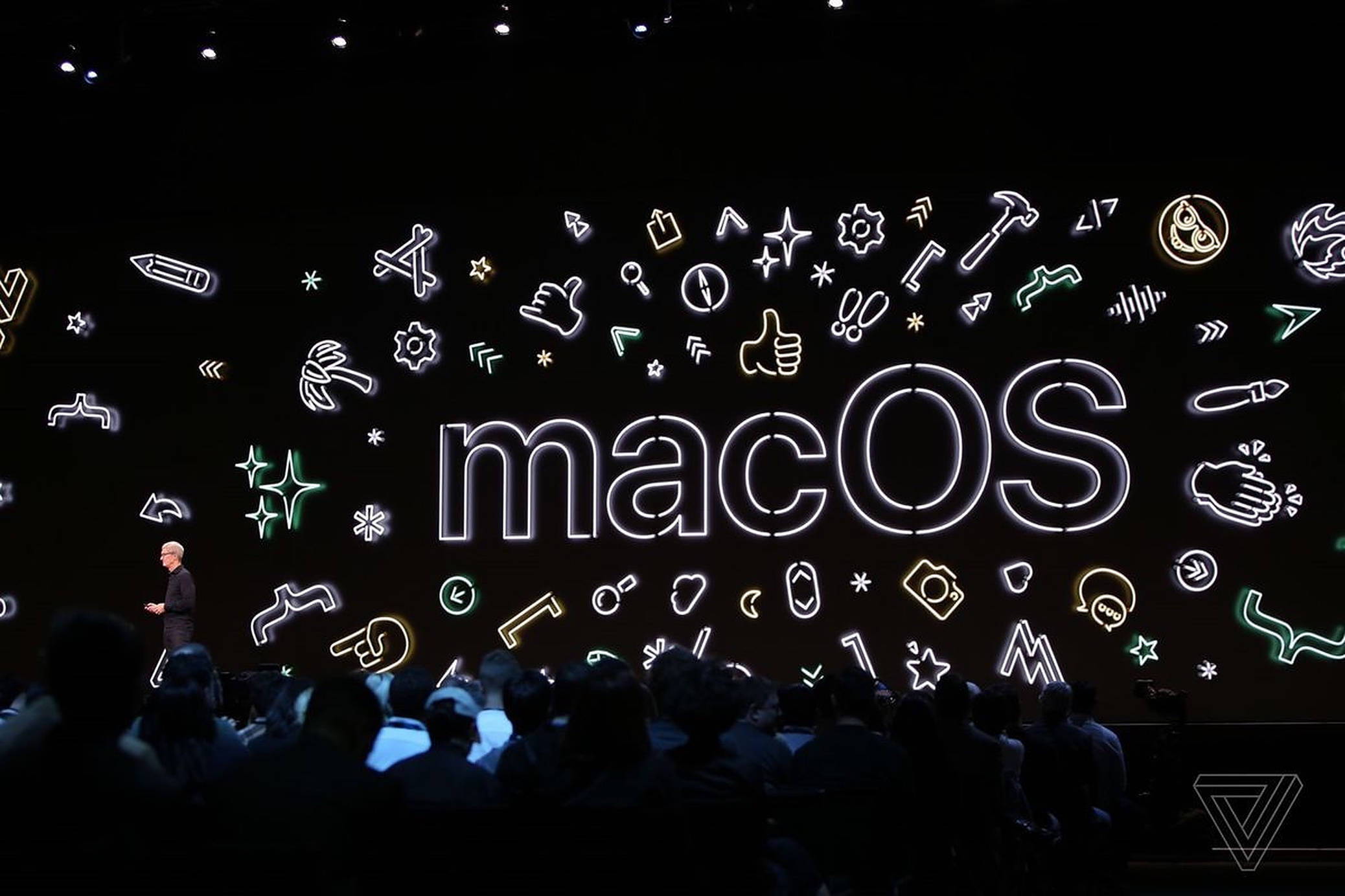 macOS Catalina 桌布 壁紙 — 演講者在黑暗舞台上展示，帶有發光霓虹 macOS 標誌和工具圖標，背景為黑色。