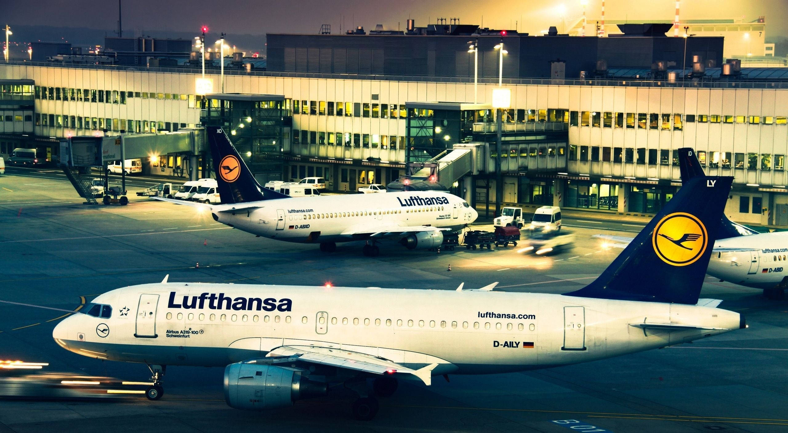 飛機 桌布 壁紙 — 漢莎航空飛機在溫暖的晚間燈光下停泊在機場航廈，地面車輛造成動態模糊。