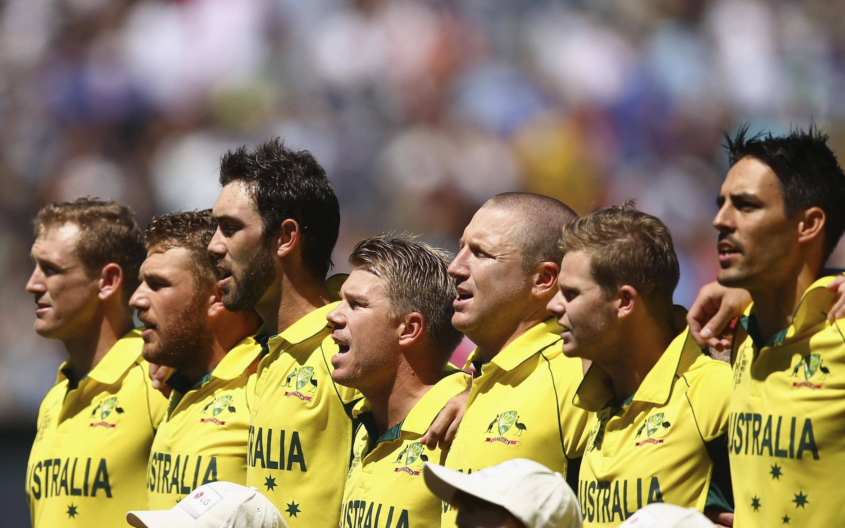 australia cricket 桌布 壁紙 — 澳洲板球隊身穿黃色球衣在體育場唱國歌，背景是模糊的人群。