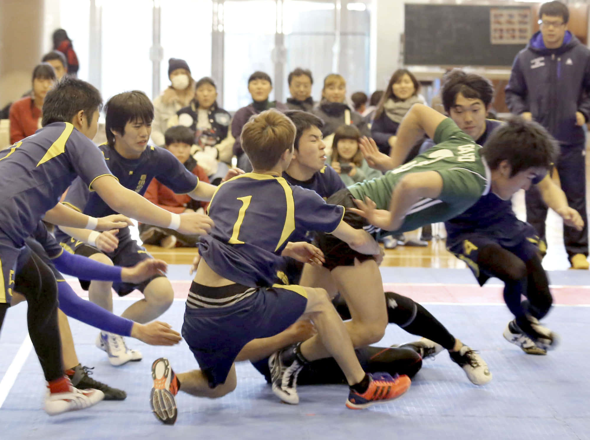 Kabaddi 桌布 壁紙 — 激烈的室內橄欖球比賽，球員身穿藍色和綠色球衣，在藍色墊子上激烈纏鬥，觀眾在場邊觀看。
