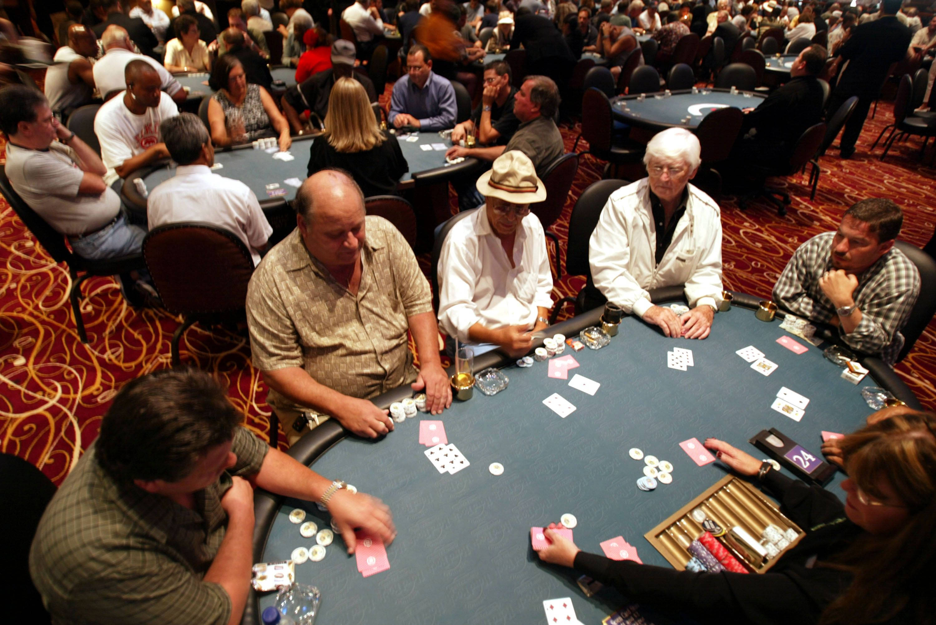 Poker 桌布 壁紙 — 激烈的撲克玩家在擠滿人的賭場房間裡，圍著一張綠色毛氈桌，地上鋪著紅色圖案地毯，全神貫注。