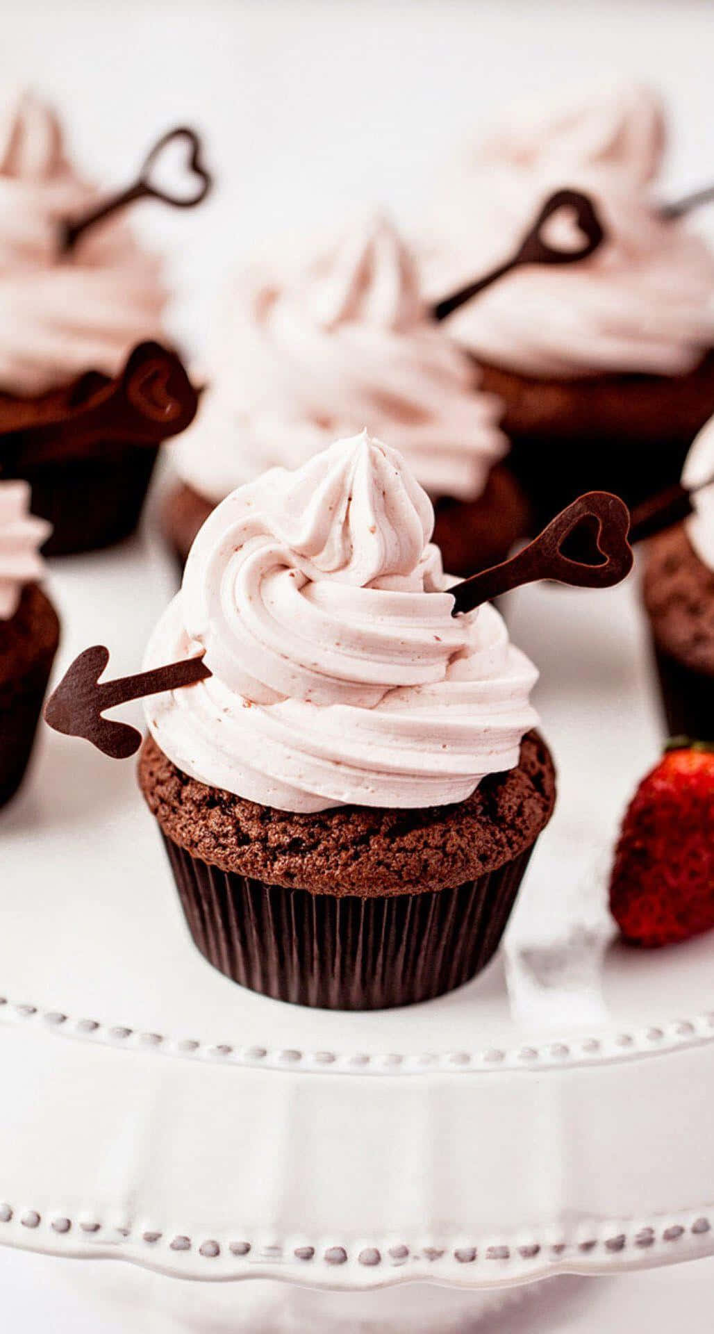 Cupcake 桌布 壁紙 — 濃郁的巧克力紙杯蛋糕，上面有粉紅色糖霜和心形巧克力鑰匙，放在白色蛋糕架上，旁邊有新鮮草莓。