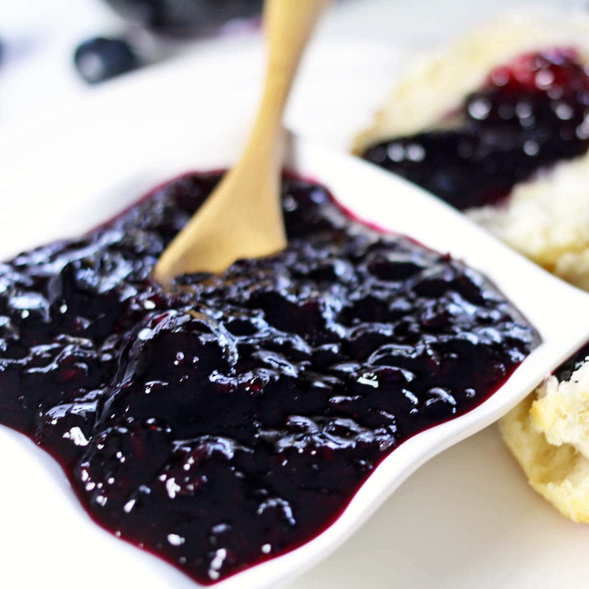 Blueberry Jam 桌布 壁紙 — 濃郁的深色藍莓果醬在白色盤子中閃閃發光，配以木勺，旁邊是金黃色的司康餅，上面有一勺果醬。