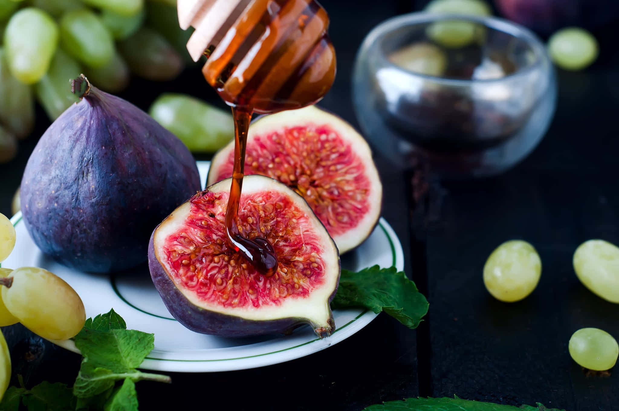 Purple Figs 桌布 壁紙 — 濃郁的蜂蜜滴落在深色表面上的切片無花果上，周圍環繞著葡萄和薄荷。